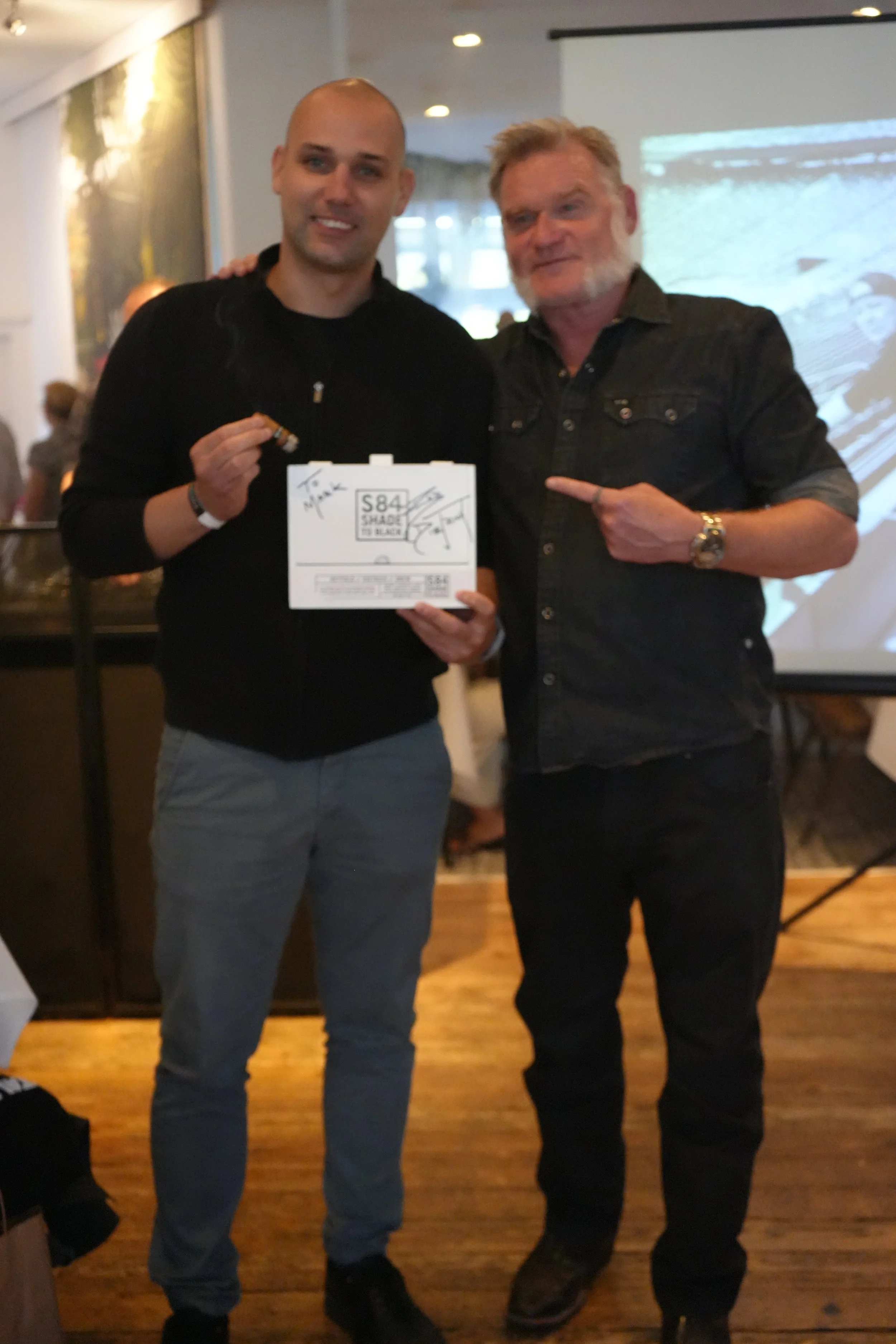Two men standing next to each other indoors, one holding a small box and the other pointing at it, with a projection screen in the background.
