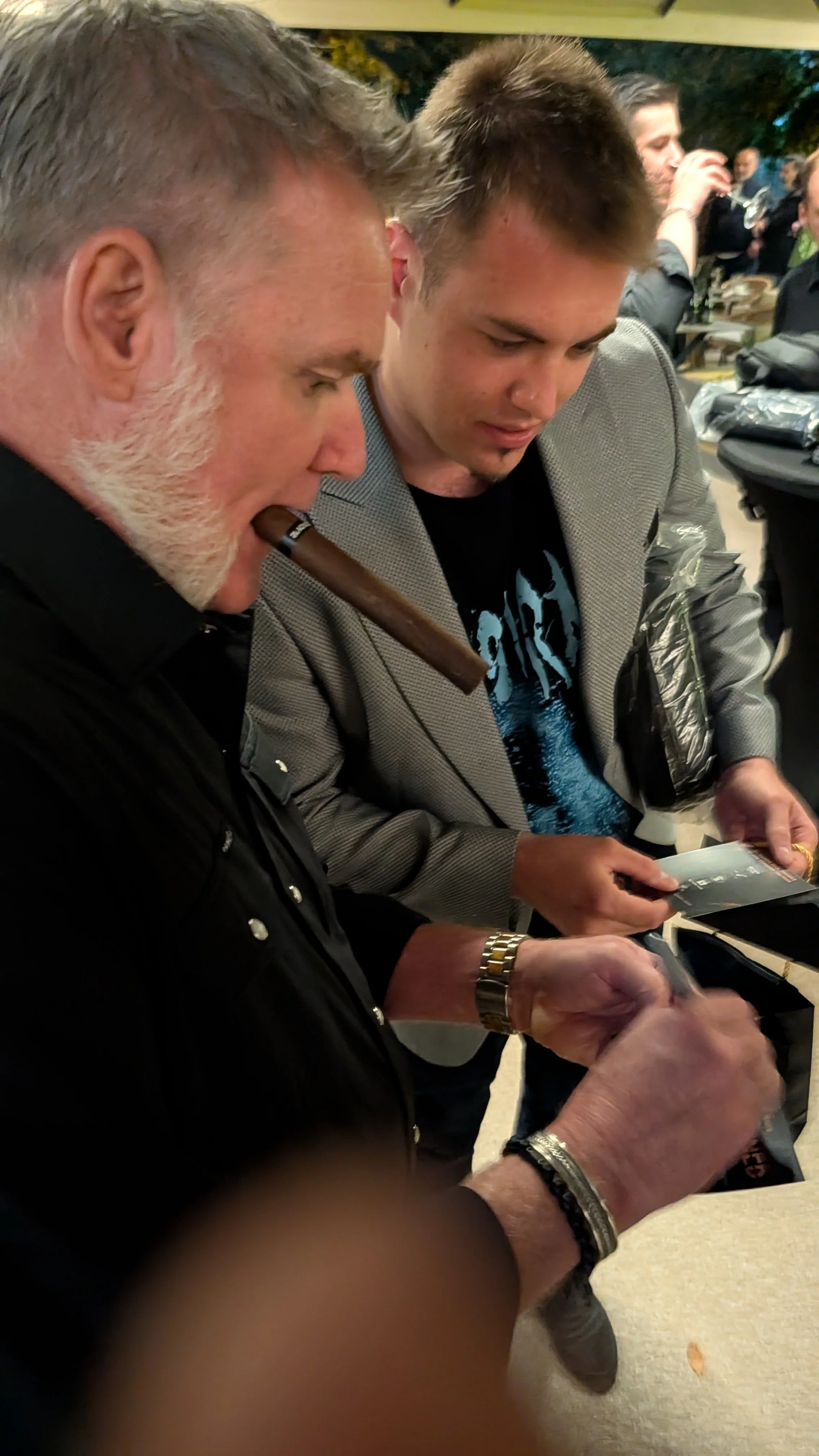 Two men are closely examining items on a table at a social gathering, with other people in the background, some holding drinks.