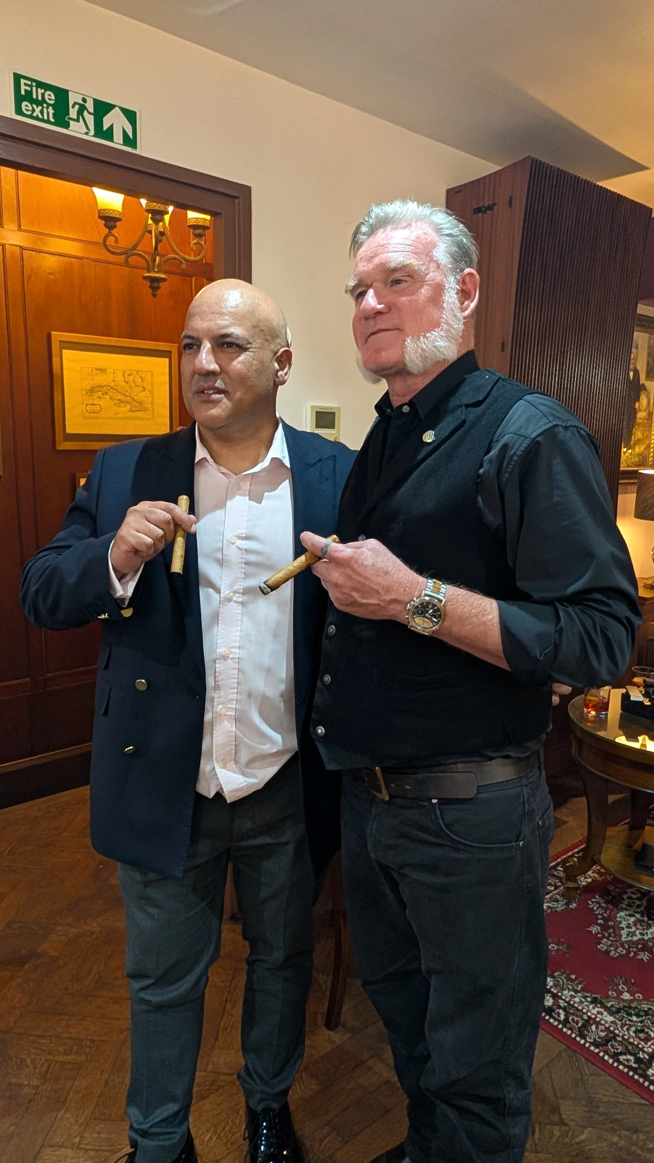 Two men holding cigars, standing indoors with warm lighting, a wooden wall with paintings, and a patterned rug in the background.