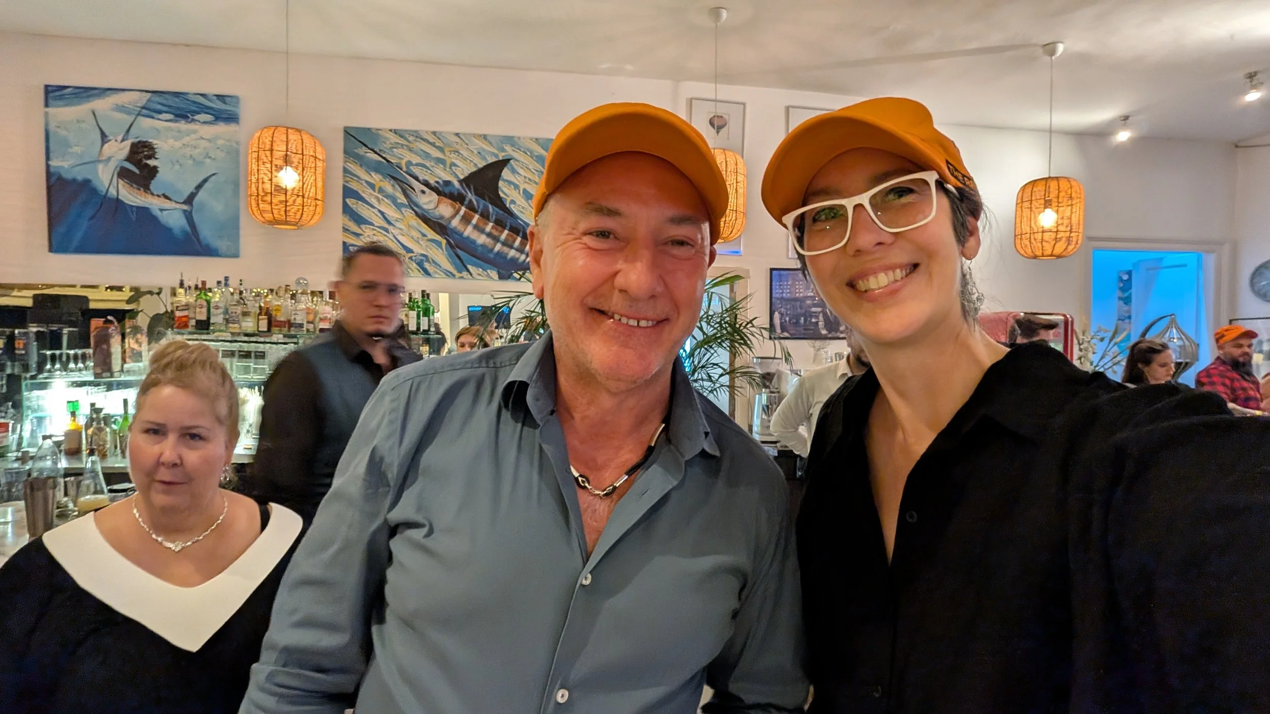 Two smiling people wearing yellow hats taking a selfie in a restaurant, with a bar, colorful paintings, and other patrons in the background.