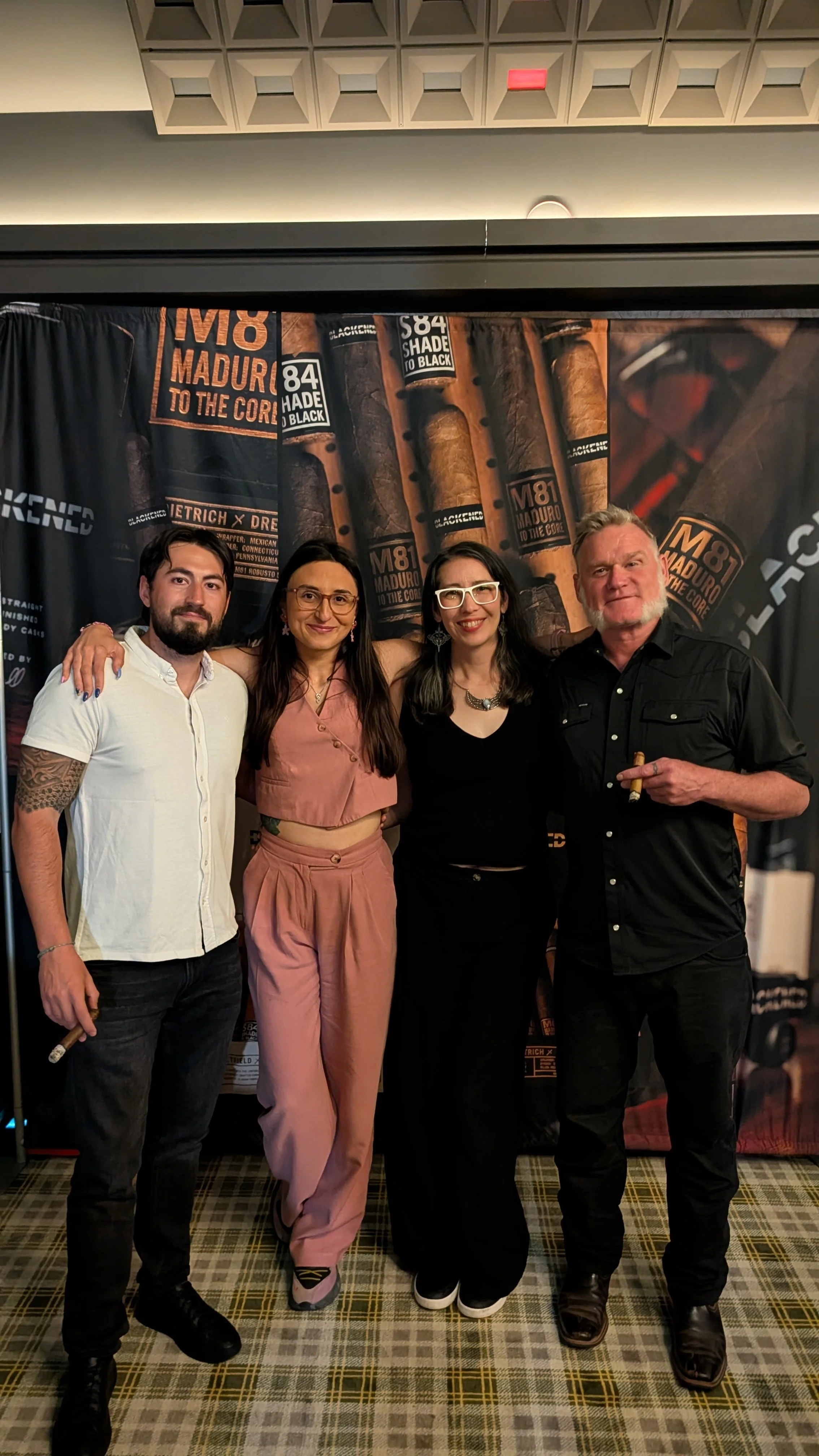 Four people standing together at an event with a cigar-themed backdrop, smiling for the photo.