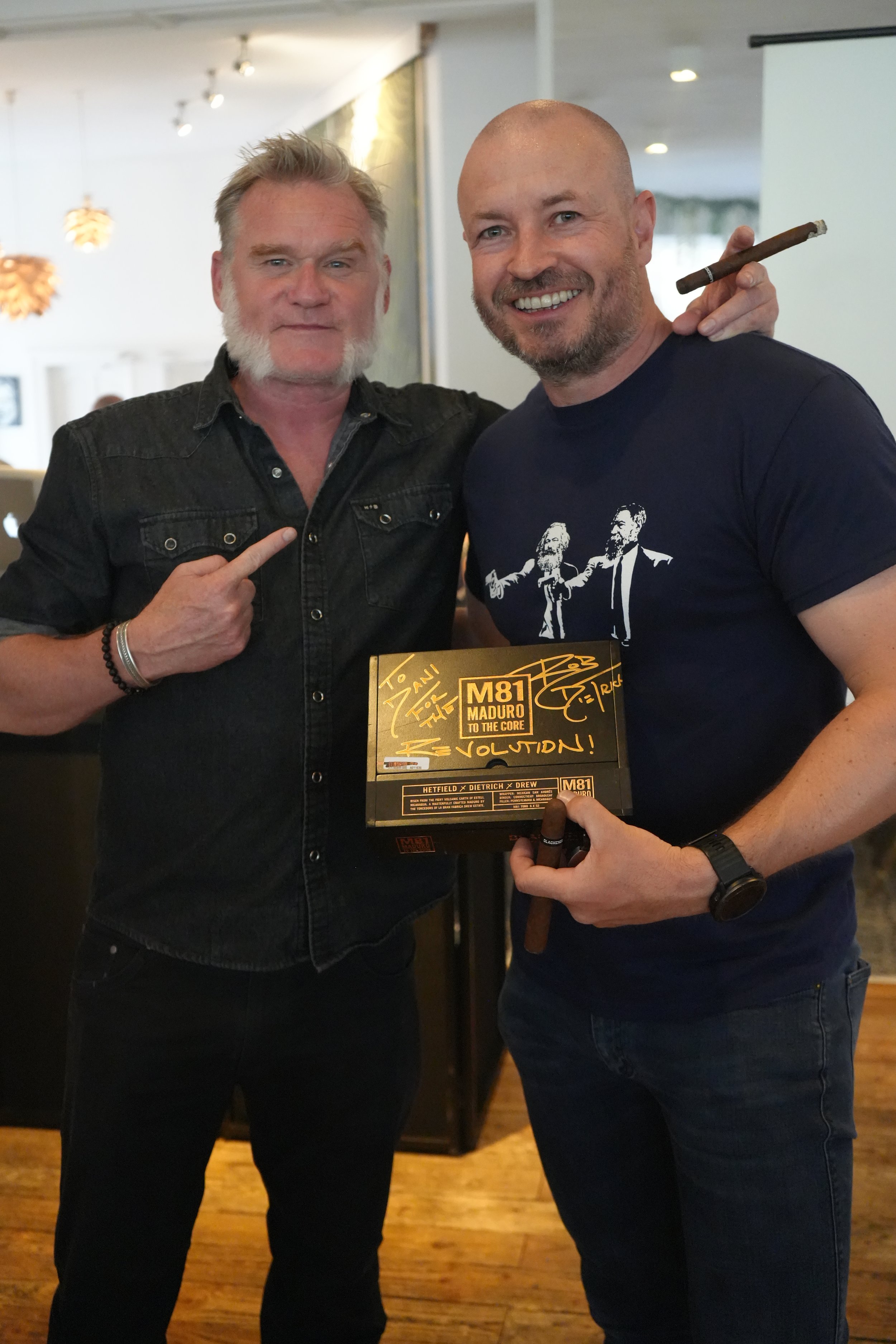 Two men standing together smiling, one holding a signed award plaque, and the other pointing at him. The man on the right has a cigar on his shoulder and is wearing a black T-shirt with graphics. The man on the left has a beard, wearing a dark button