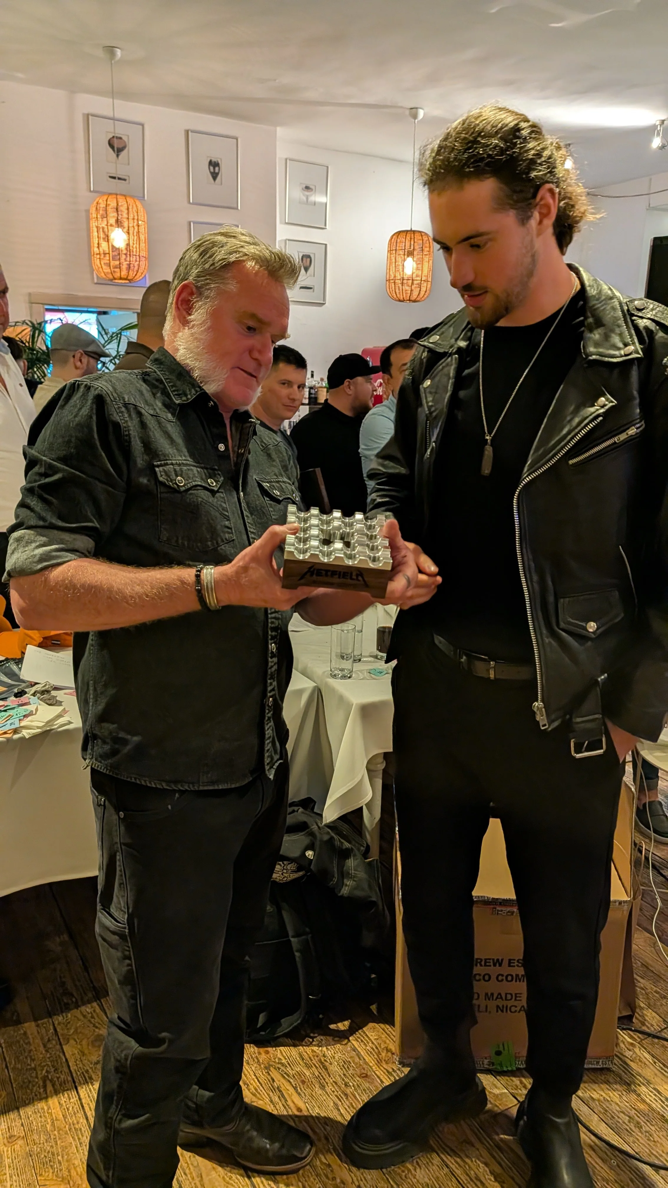 Two men are looking at an object in an indoor setting with several people in the background, and decorative lighting on the ceiling.