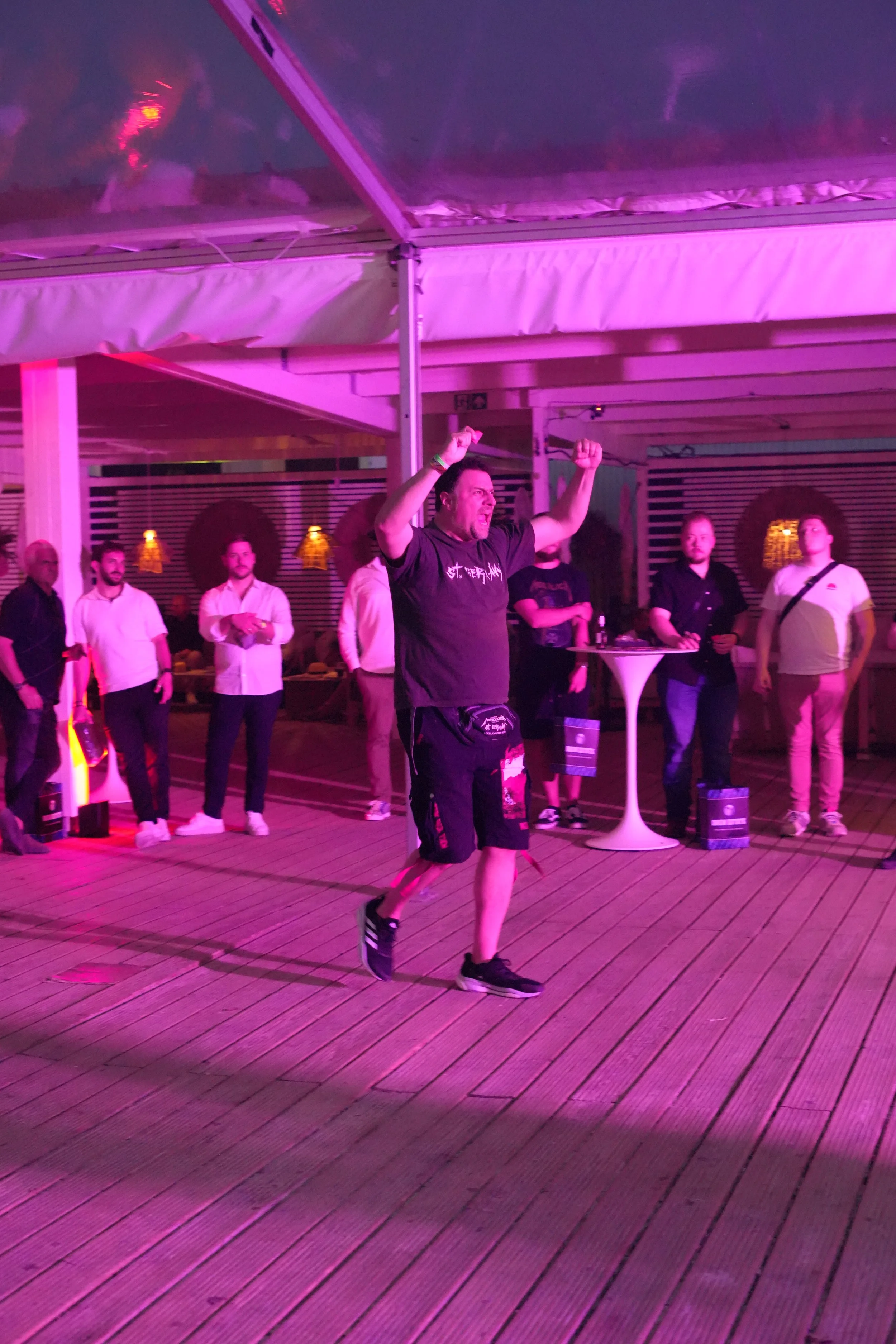 A man in black shirt and shorts running on a wooden deck at night, with a group of people watching, under purple lighting.