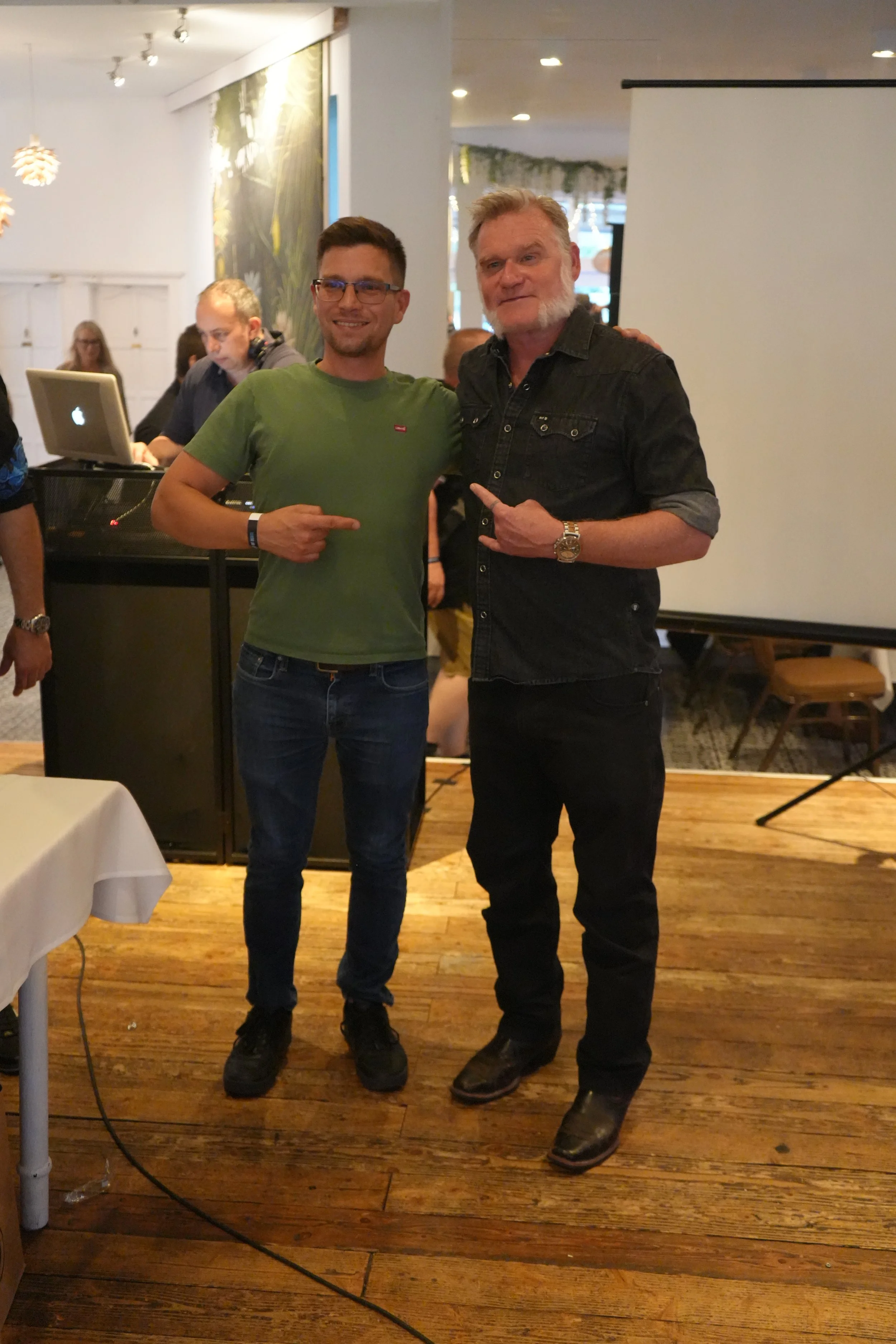 Two men are standing in a room, smiling and pointing at each other. The man on the left is wearing glasses, a green t-shirt, and dark jeans. The man on the right has gray hair and a beard, dressed in a black denim shirt and dark pants. In the backgro