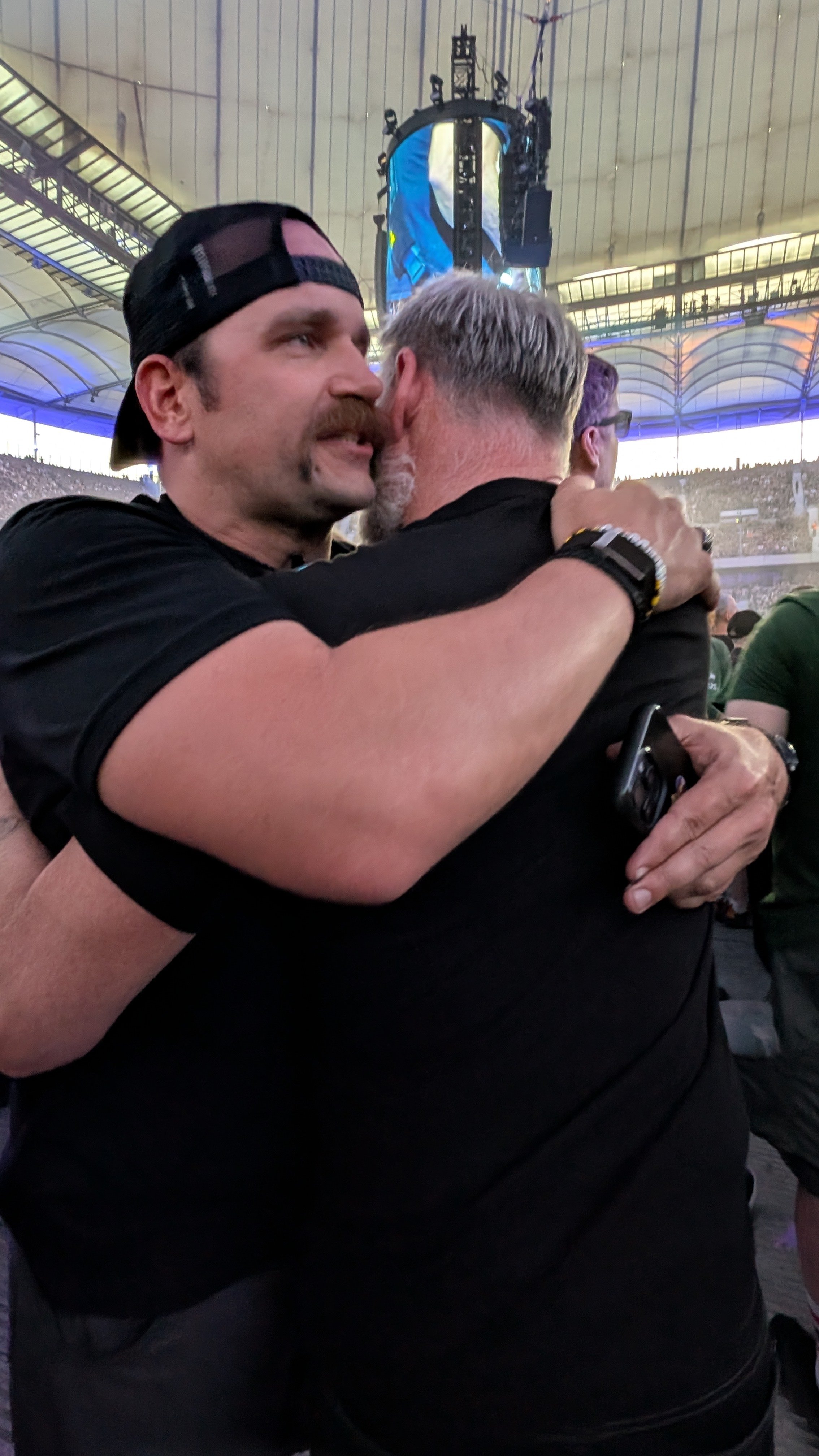 Two men embracing at a crowded event or concert. One has a black baseball cap worn backward, mustache, and beard, while the other has gray hair and a beard. The background shows a large indoor stadium with a high, arched ceiling, stage, and screens.