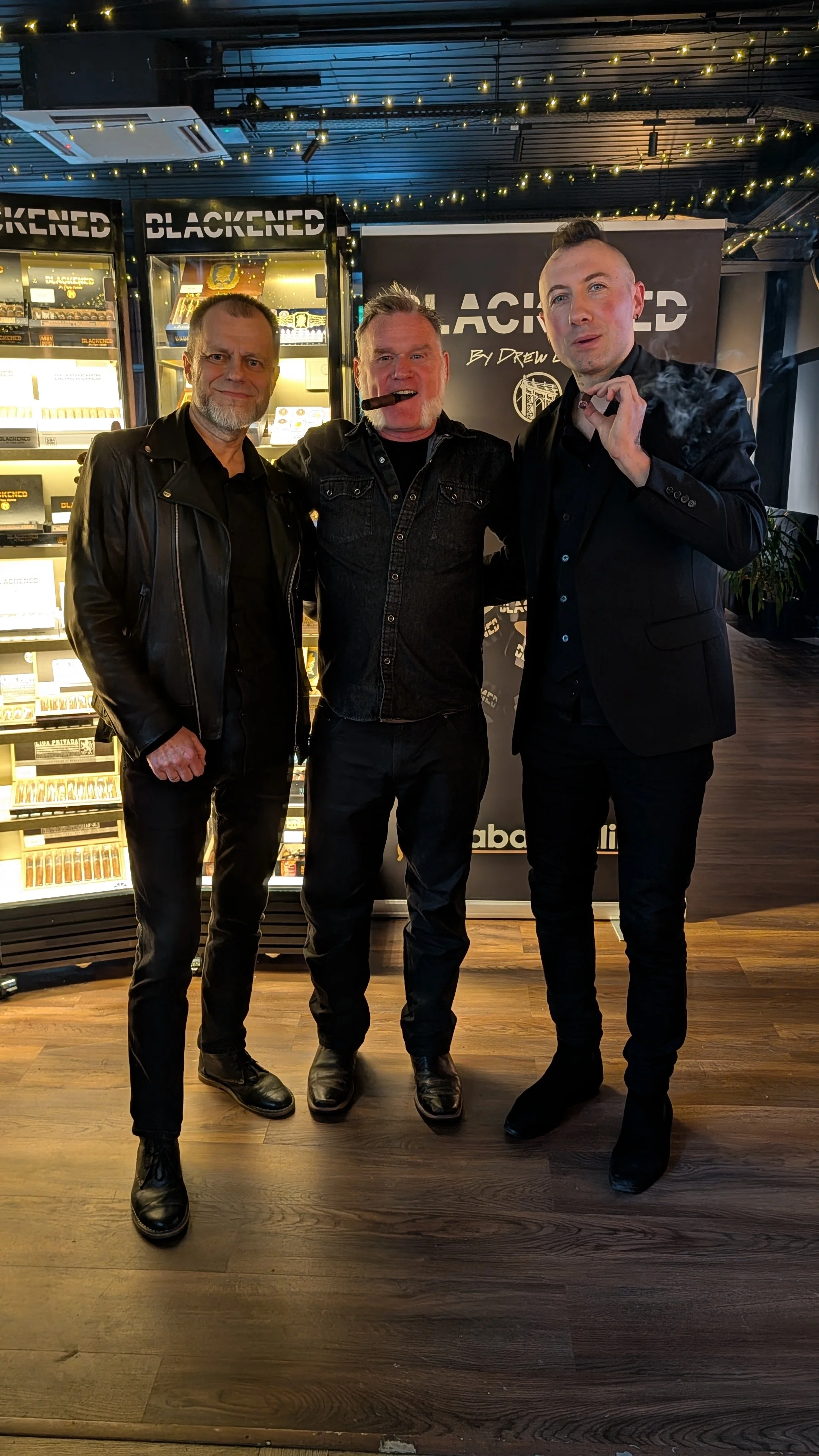 Three men standing together at an event, with a black backdrop that reads 'BLACKENED' by Drew. The man in the middle is smoking a cigar. They are dressed in dark clothing, and behind them are illuminated display cases with cigars and a banner with th