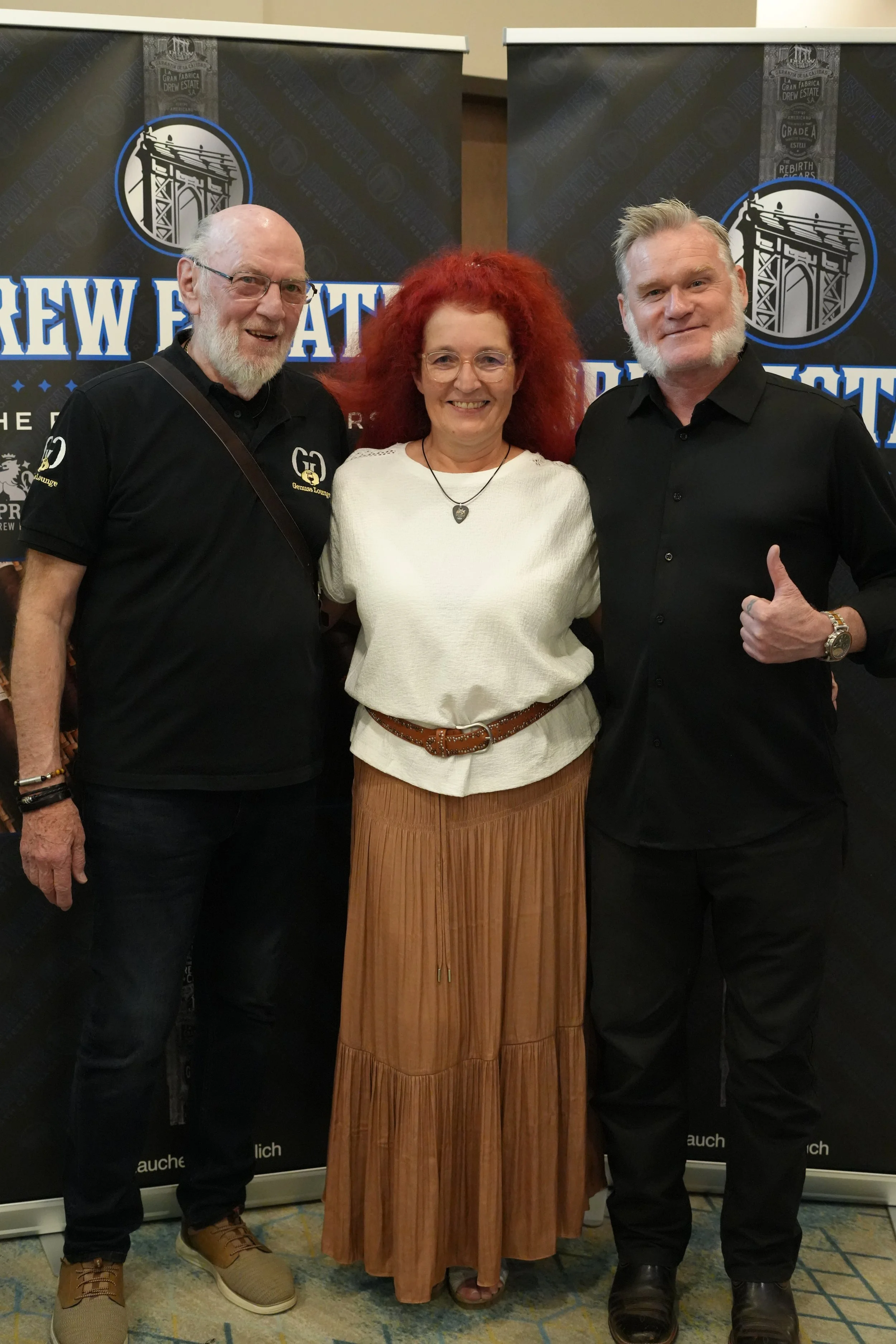 Three people standing in front of a backdrop with the words "brew" and "weich" visible. The person on the left is an elderly man with glasses, a beard, and a black shirt with embroidered logo, smiling. The person in the middle is a woman with curly r