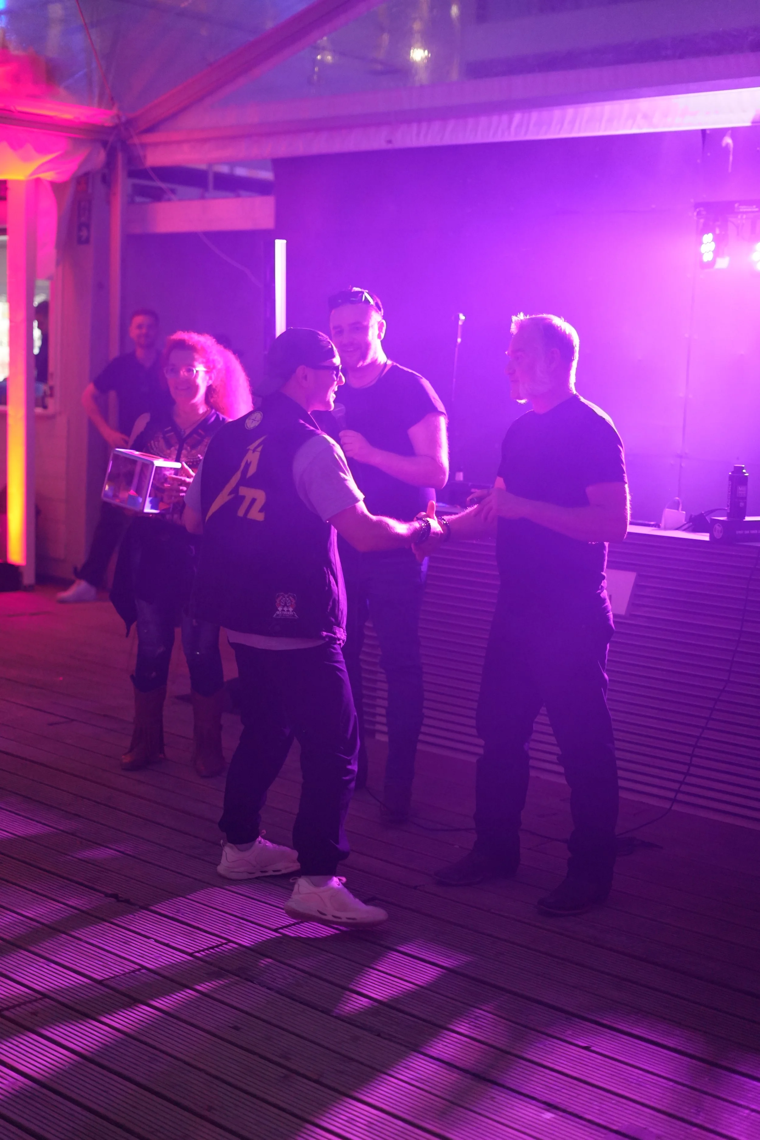 A group of people socializing at a party or nightclub under purple and pink lighting, with some shaking hands and others engaging in conversations.