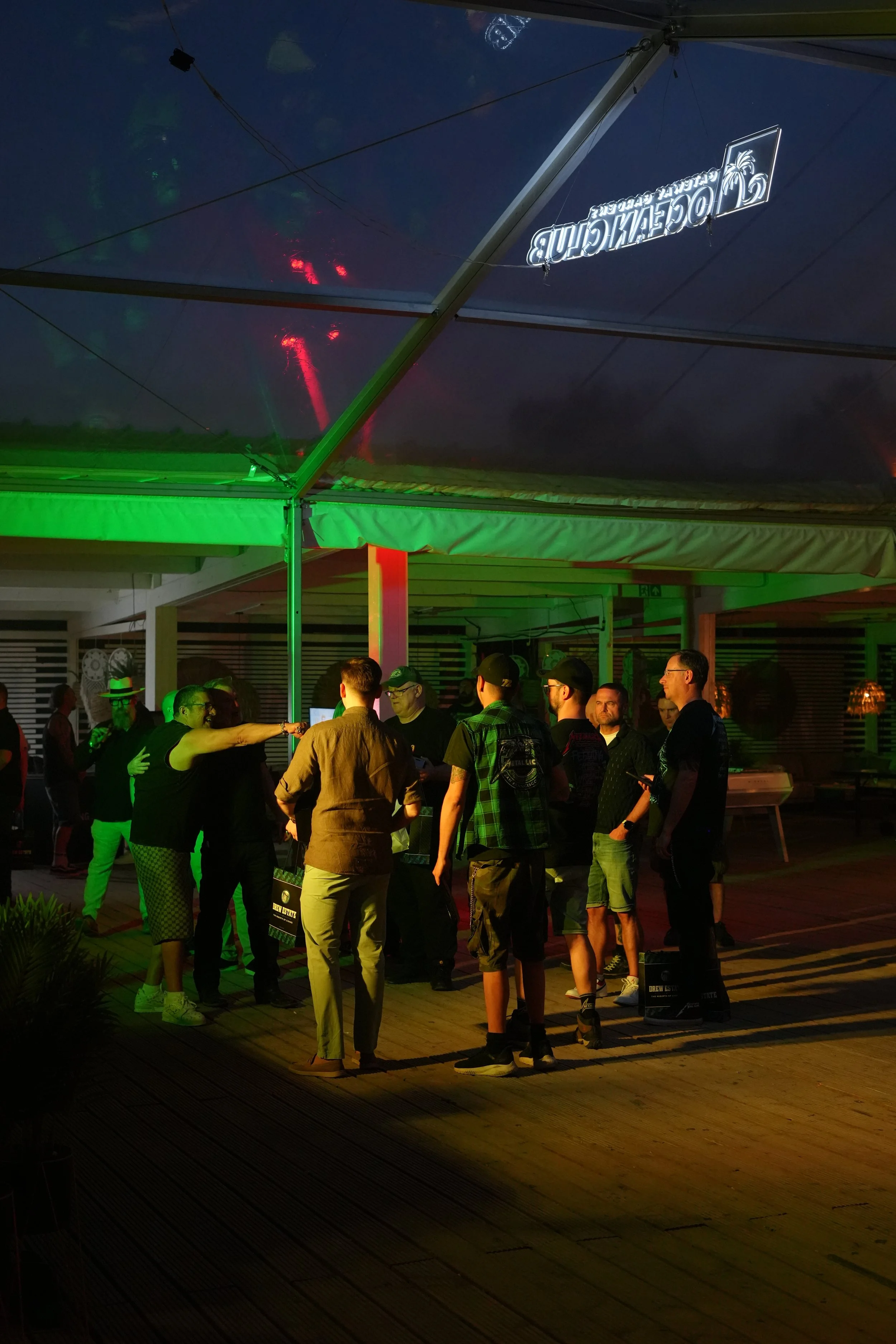 A group of people gathered under a patio with a clear roof, illuminated by green lighting, during nighttime. Some people are talking and holding items, and a man with a wheelchair is visible among the crowd.