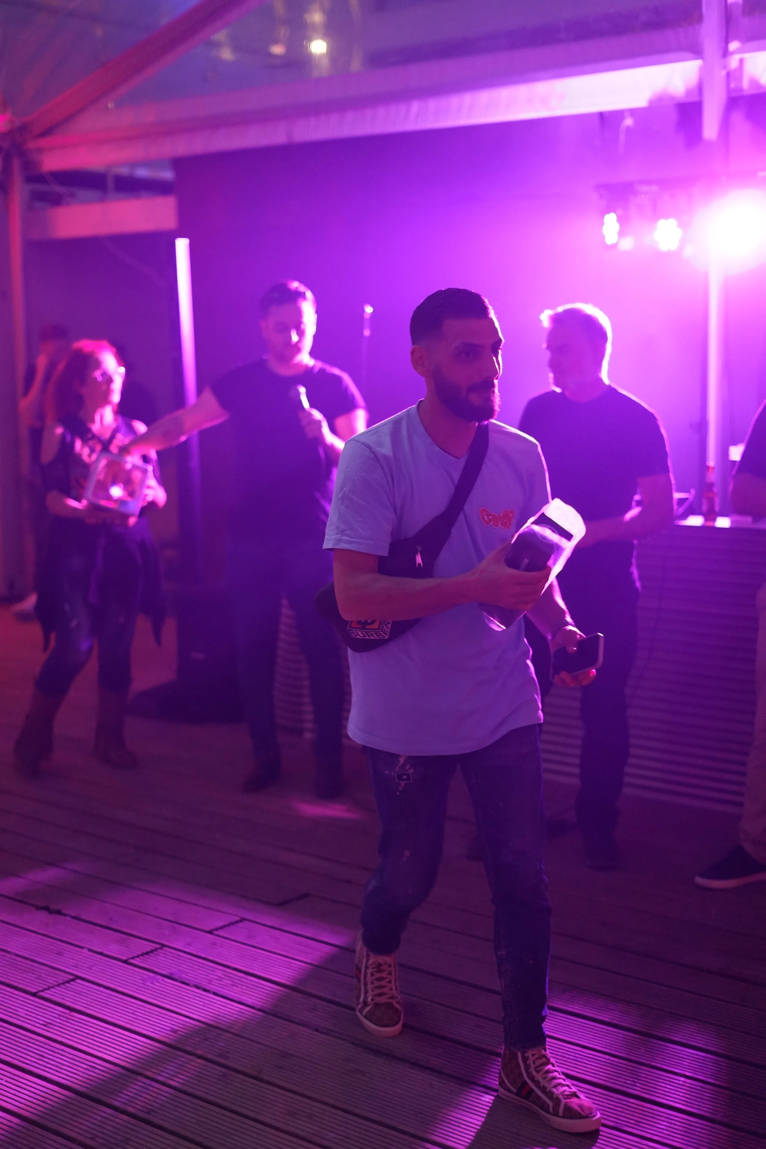 A group of people in a dimly lit room with purple lighting, standing and walking on a wooden floor. One man in the foreground is holding a phone and booklet, wearing a white T-shirt, jeans, and sneakers. Others are chatting, holding drinks, and some 