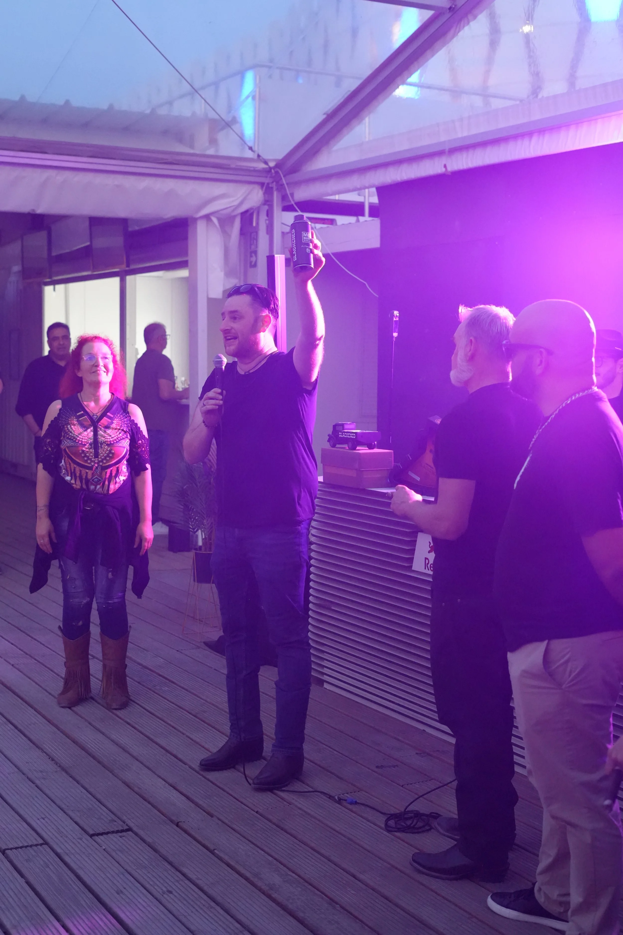 People gathered at an indoor event, with a man holding a microphone and raising an object in his hand, standing on a wooden floor, with others watching and purple lighting in the background.
