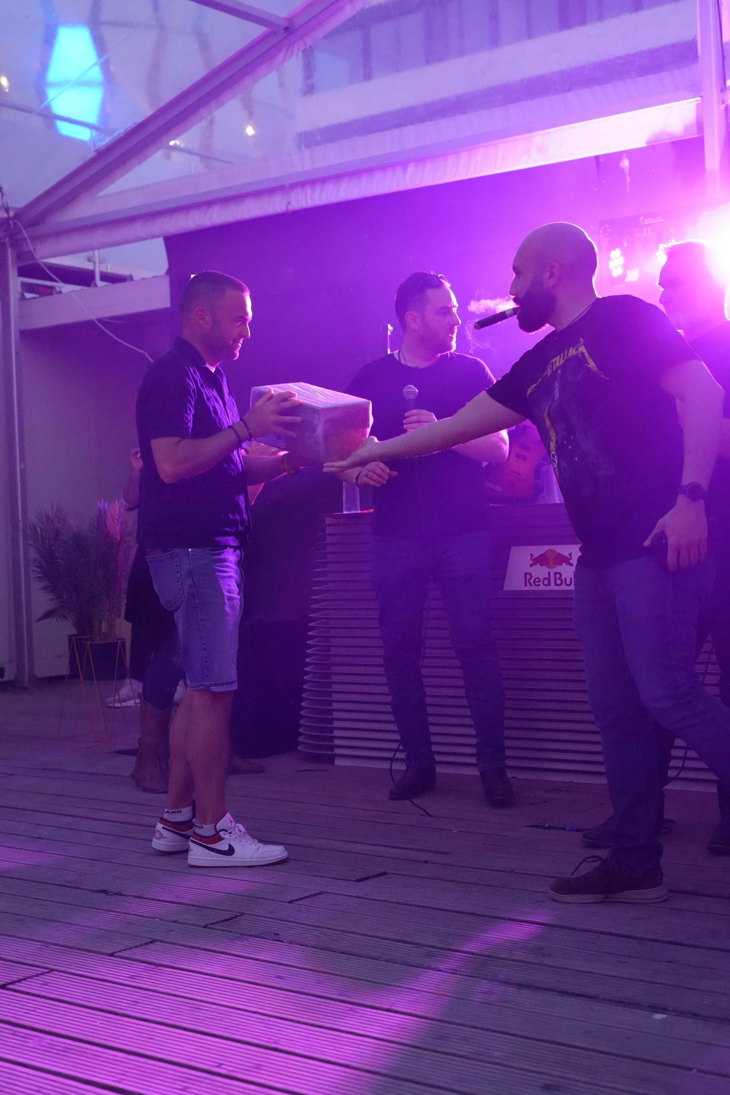 People on a stage during a nighttime event, with purple and pink lighting, one person handing a gift to another person wearing a Nike sneakers, while a woman holds a microphone.