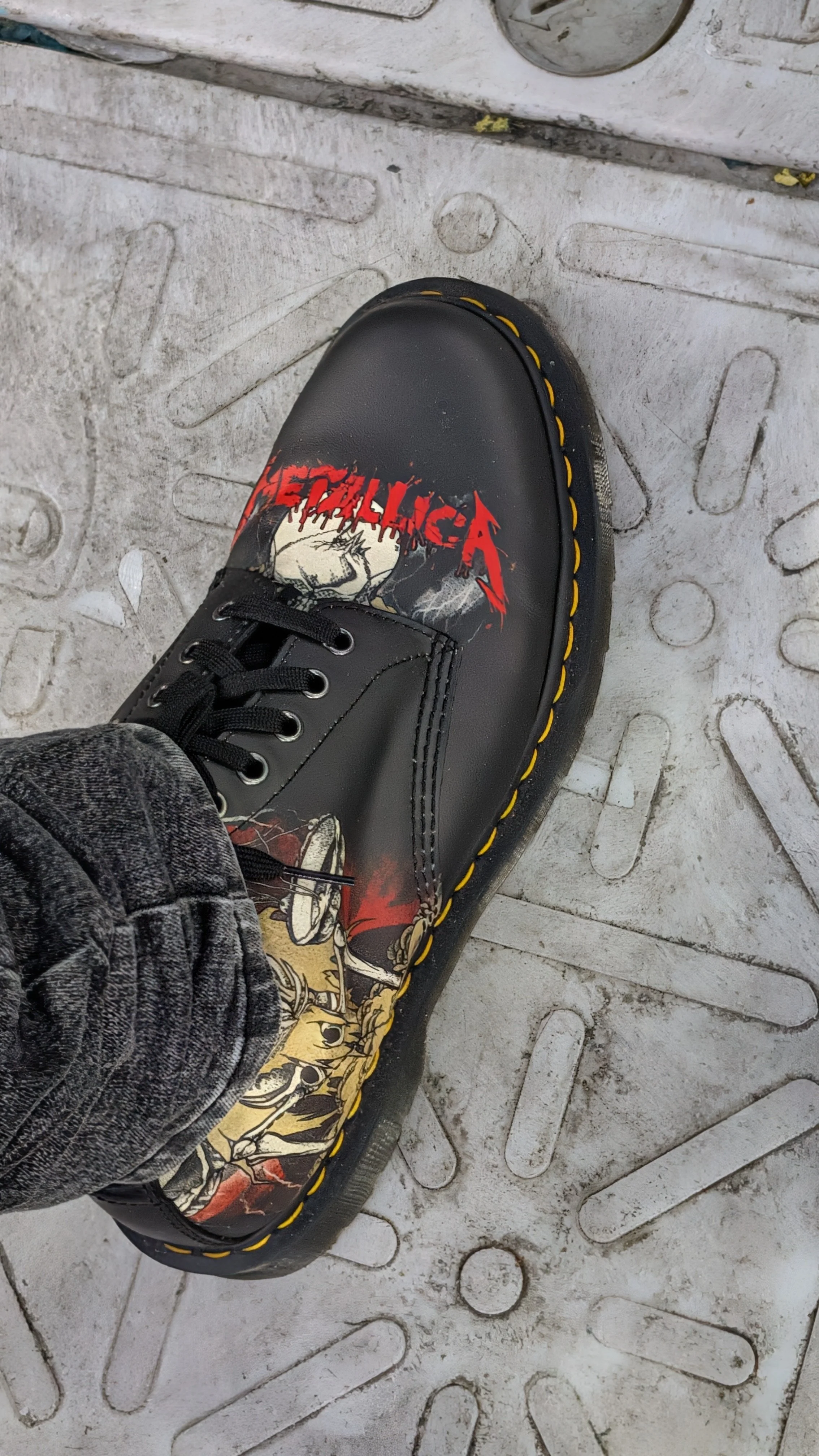 Close-up of a person's foot wearing a black leather boot with a graphic design, on a textured metal surface.