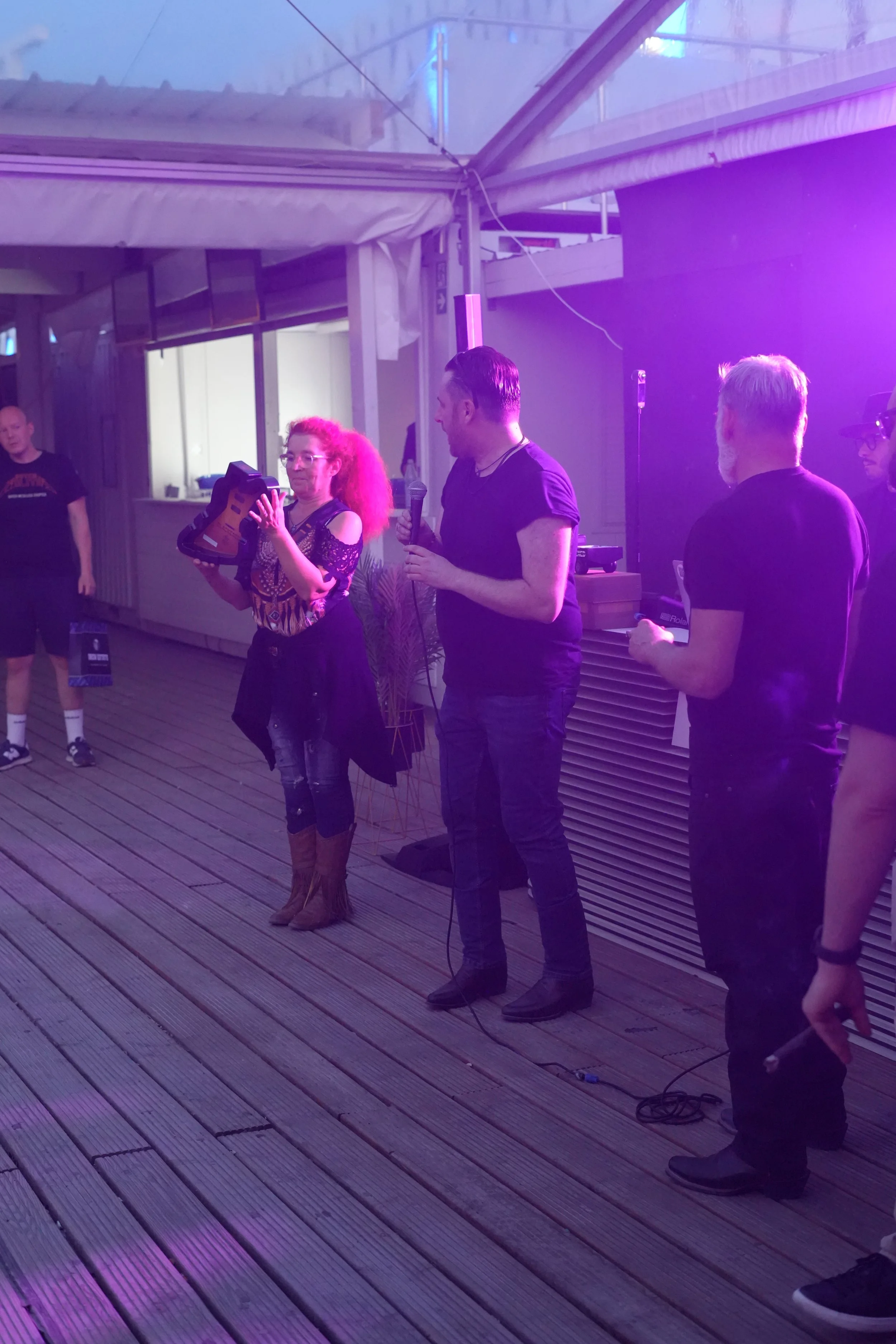 A group of people is gathered on a wooden deck with purple lighting, some are holding microphones or objects, and one woman is holding a pair of brown boots. The setting appears to be indoors or semi-outdoors, with a structure and windows in the back