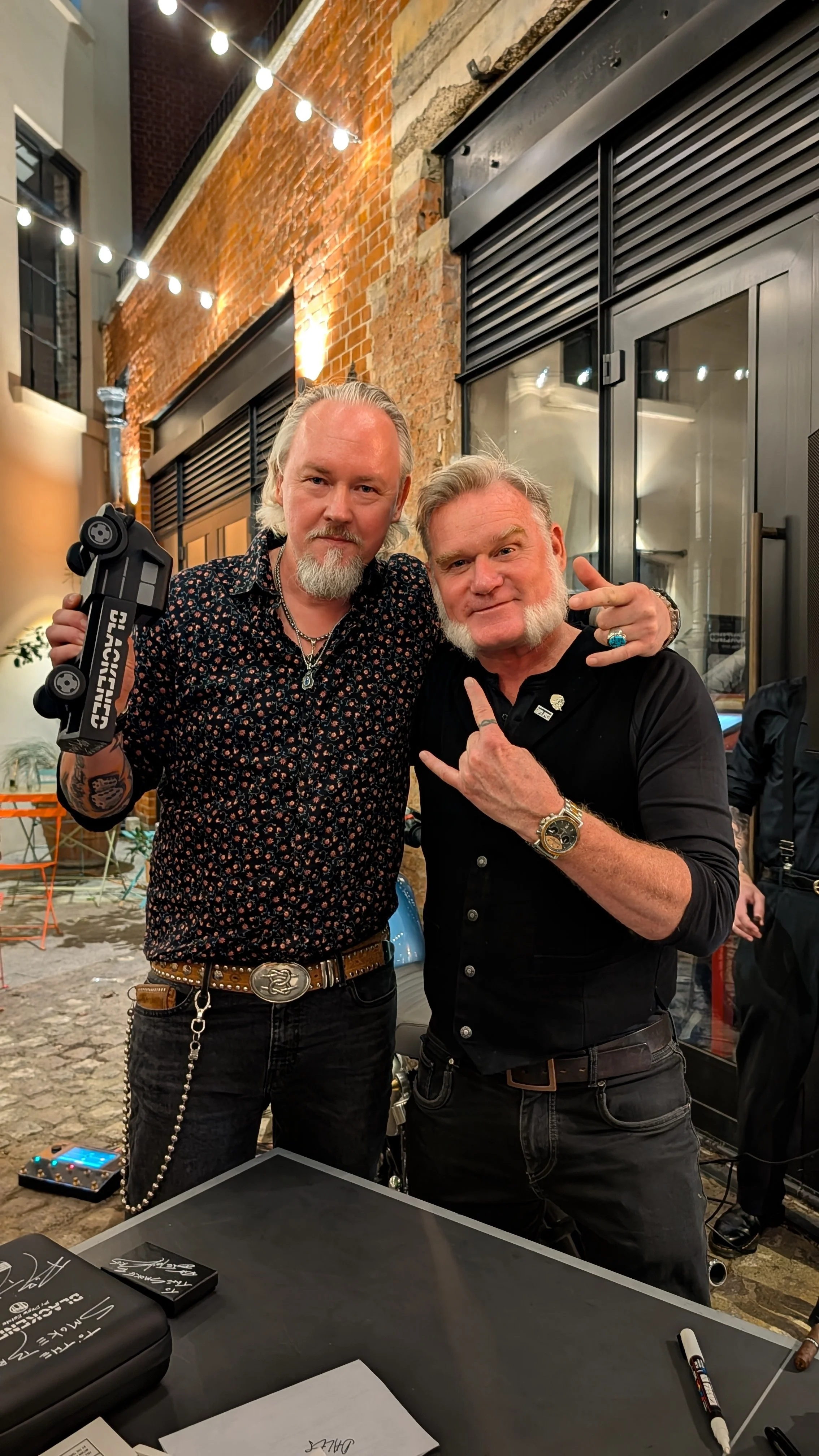 Two men with gray beards posing together at an outdoor venue with string lights, brick wall, and black metal doors in the background. One man holds a small toy car.