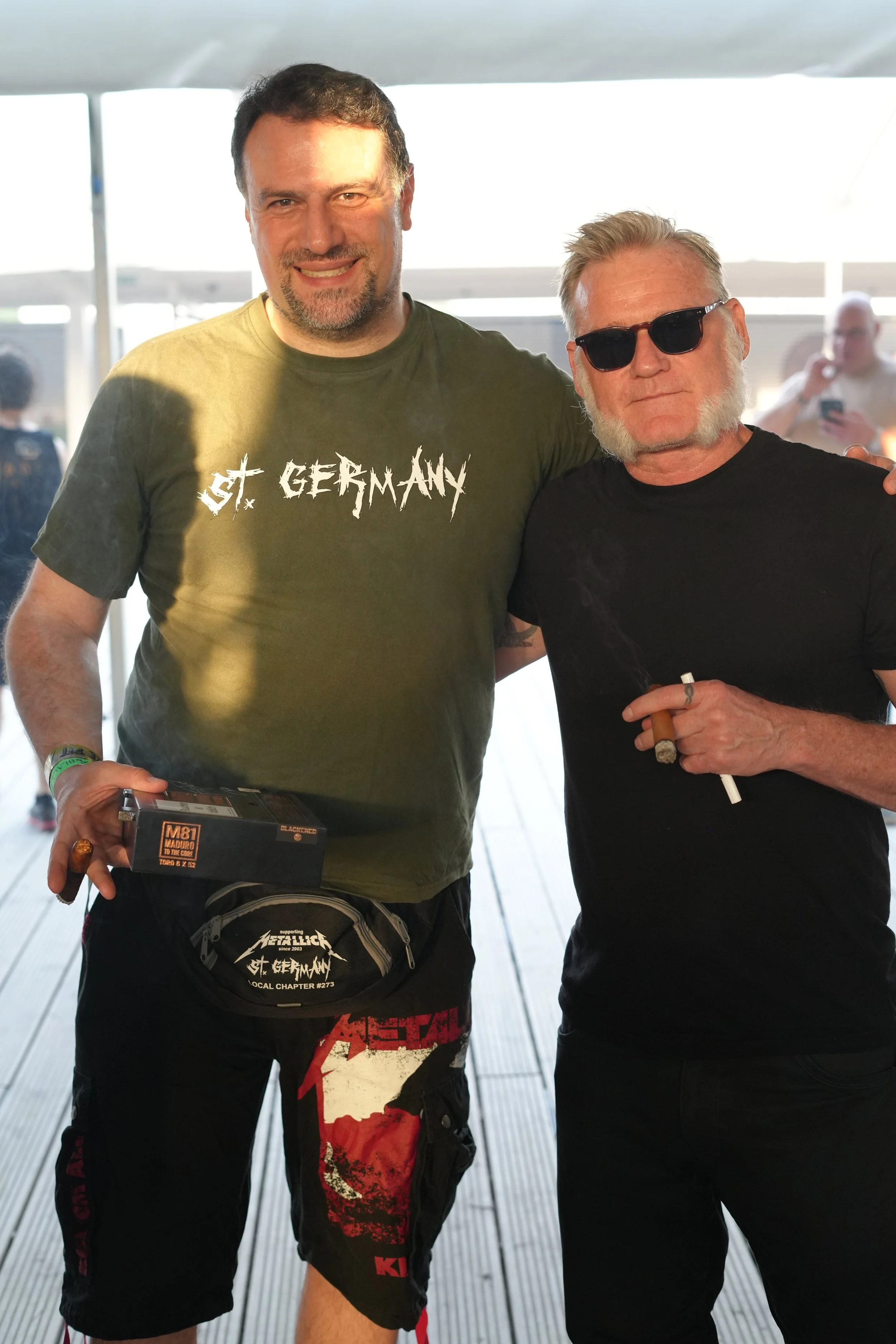 Two men standing together, one wearing a green T-shirt with 'St. Germany' text, the other wearing sunglasses and a black T-shirt, indoors with bright light in the background.