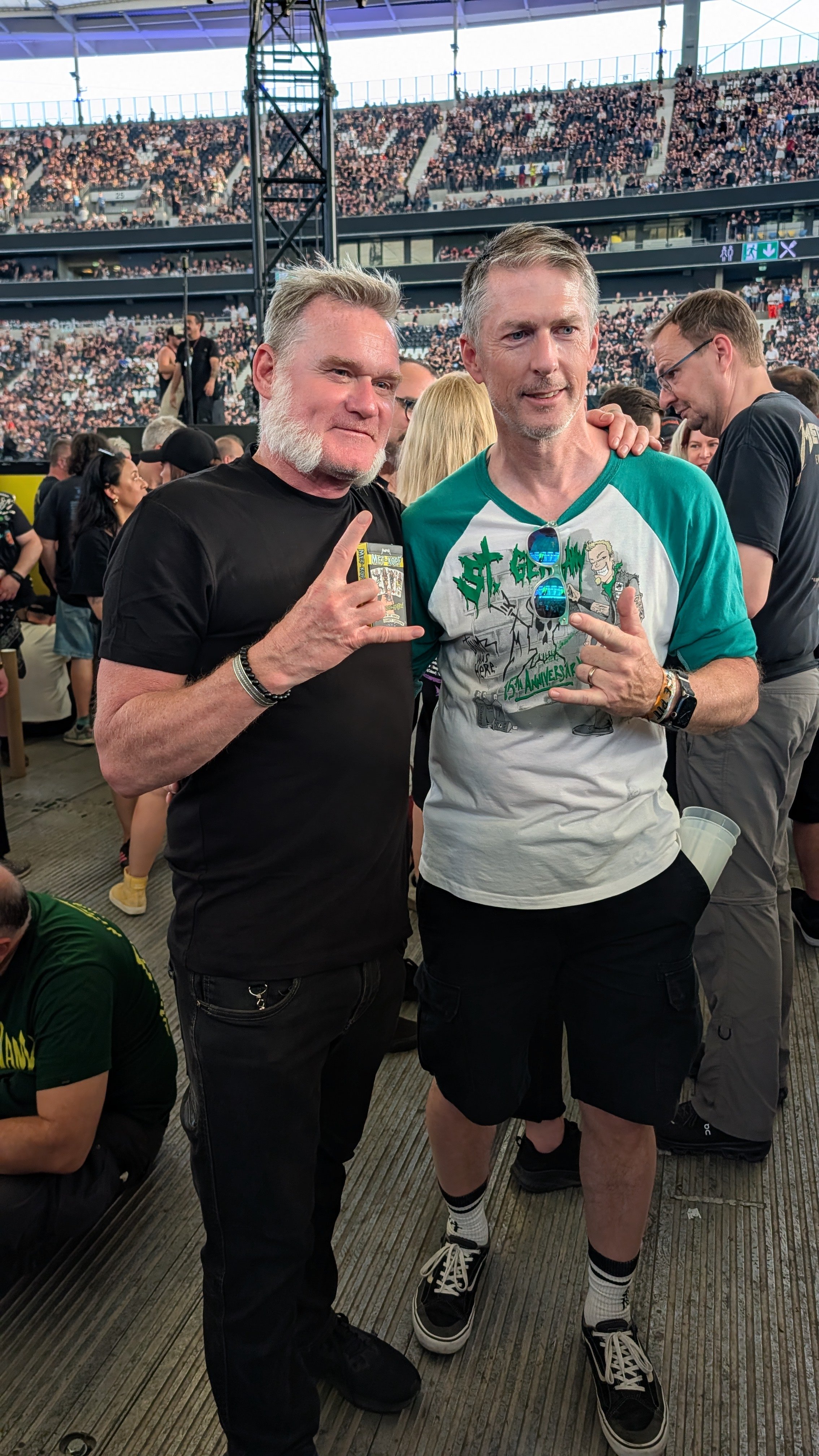Two men standing together at a crowded event, one wearing a black t-shirt and the other wearing a white and teal graphic t-shirt, with a large stadium filled with people in the background.