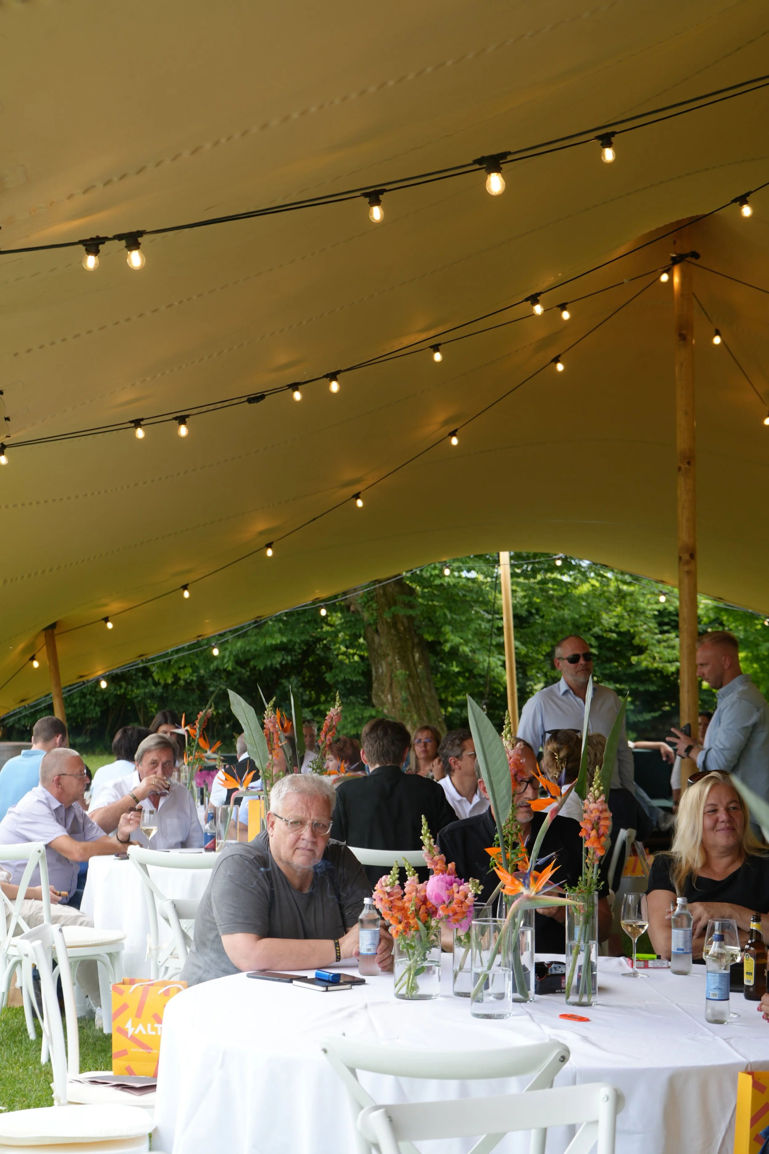 People gathered at a daytime outdoor event under a yellow canopy stringed with lights. Several tables with white tablecloths are decorated with tall, colorful floral arrangements. The guests are casually dressed, some standing and some sitting, engag