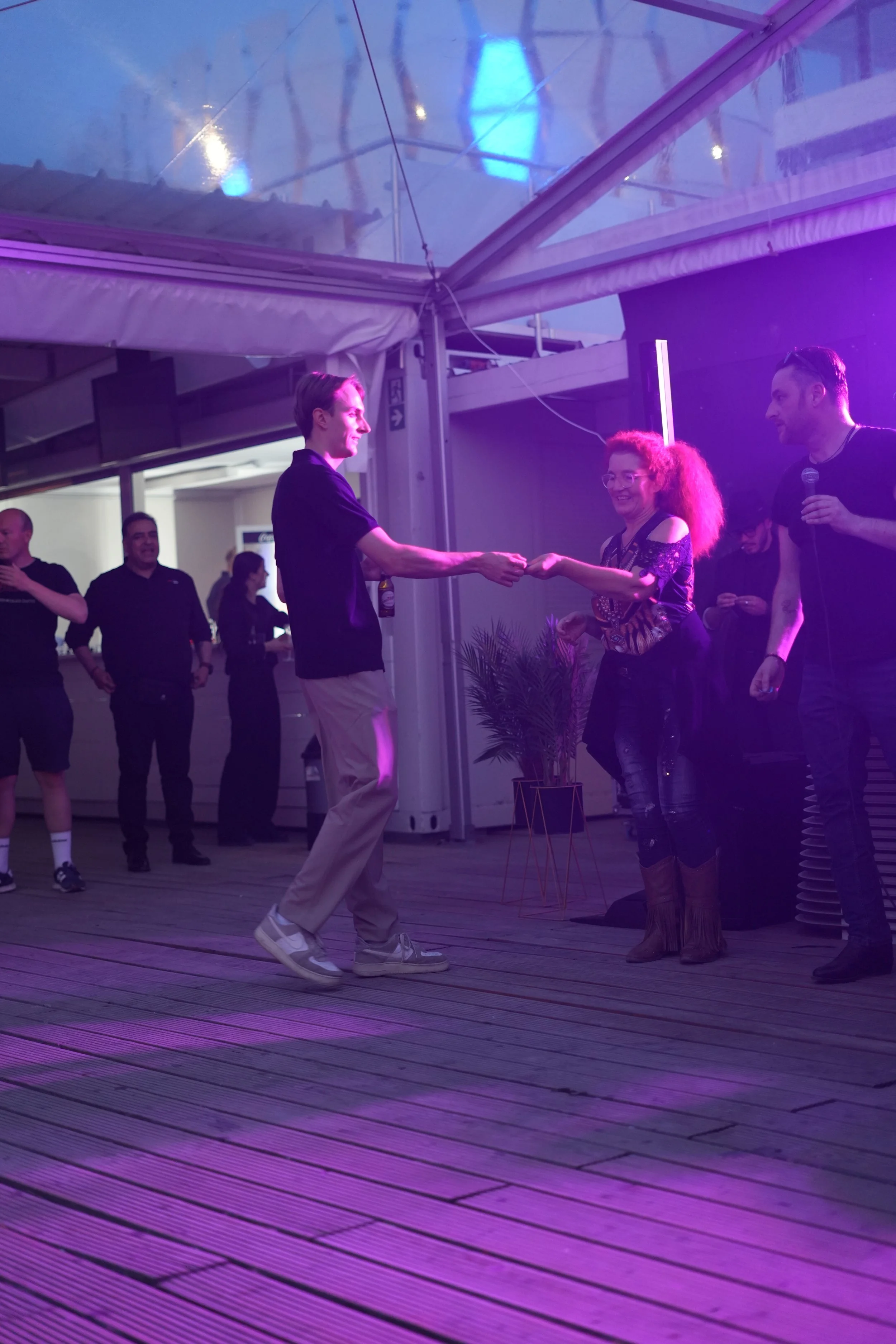 Two people are dancing together at an indoor event with purple lighting. The man on the left is wearing beige pants, a dark shirt, and sneakers, while the woman on the right has curly hair, glasses, a patterned top, and fringed boots. Several onlooke