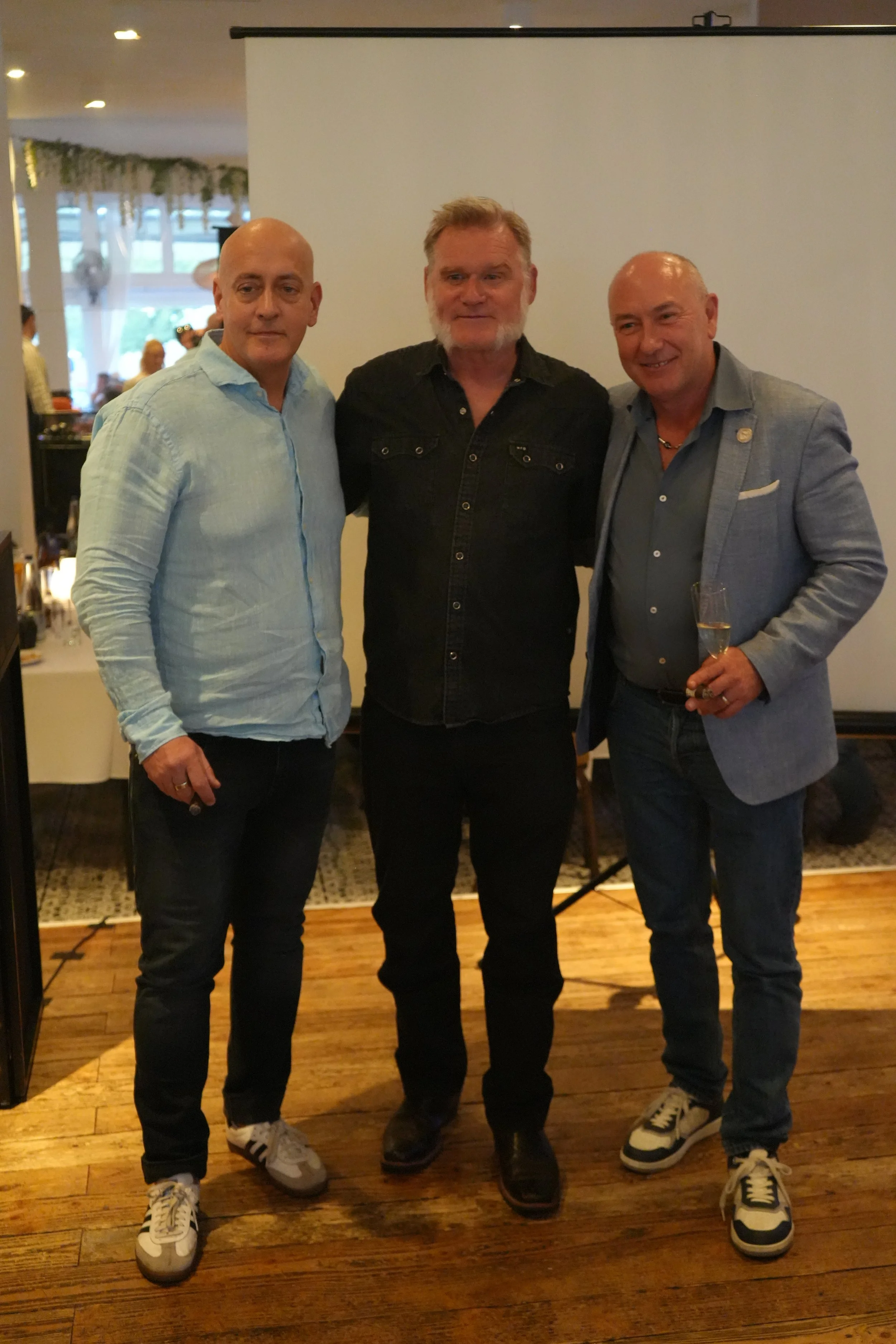 Three men standing side by side indoors in front of a blank screen. They are smiling, with one holding a champagne glass. The setting appears to be a celebration or event.
