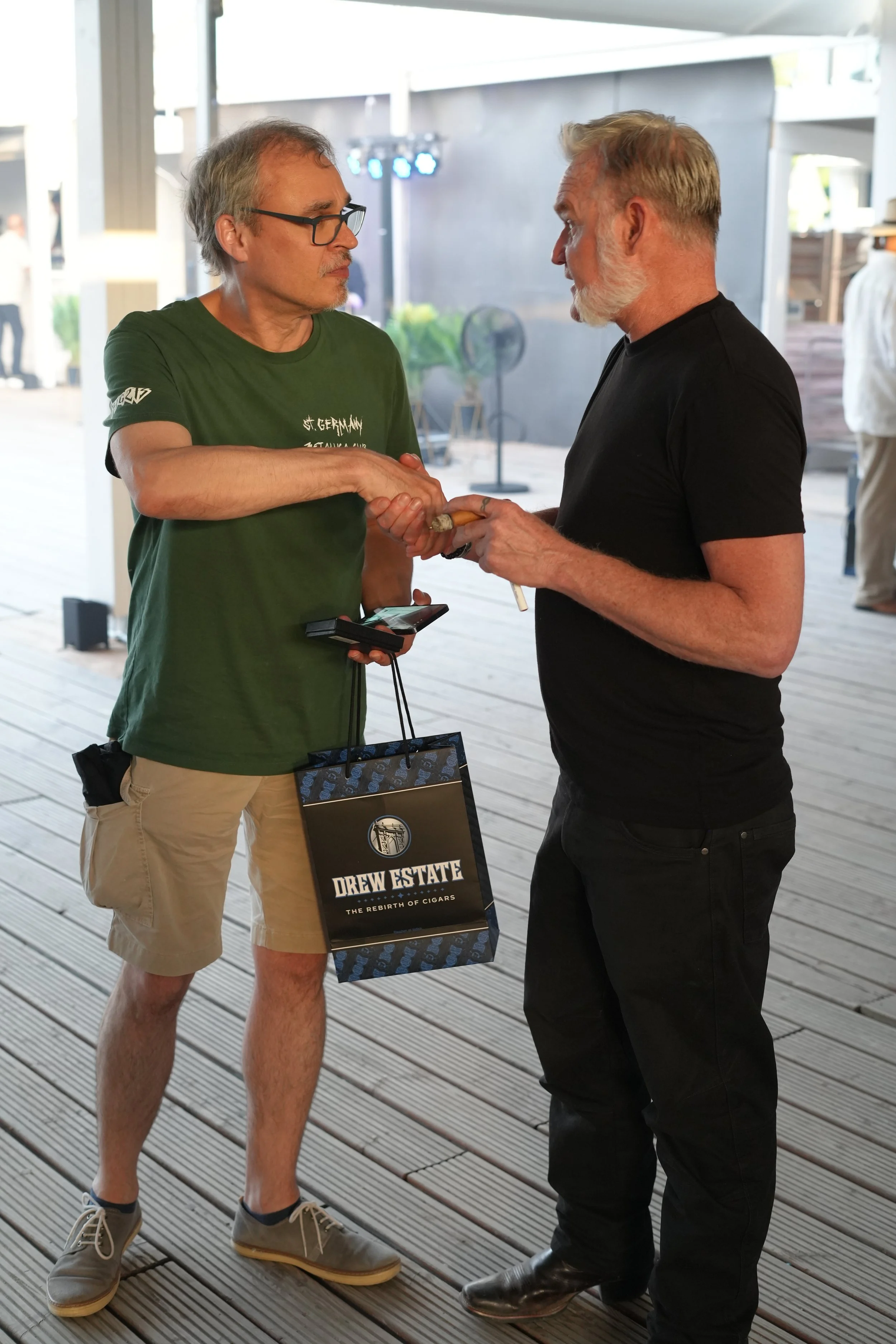 Two men shaking hands at an indoor event. One man is wearing a green T-shirt, beige shorts, and glasses, holding a smartphone and a bag with Drew Estate branding. The other man is dressed in a black T-shirt and black pants. Background shows other peo