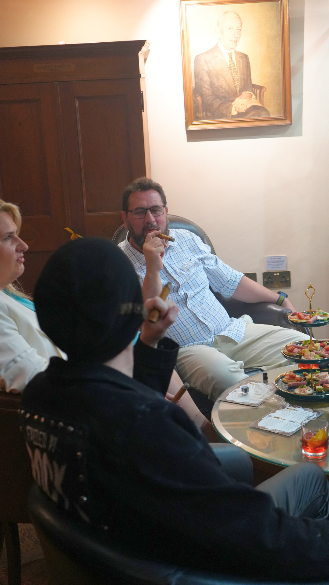 Group of people sitting in a room, holding cigars, with a man in a checked shirt sitting in a leather chair. A woman in a white blazer is in profile, and a person with a dark hoodie and black hair is in the foreground, holding a cigar. There are thre
