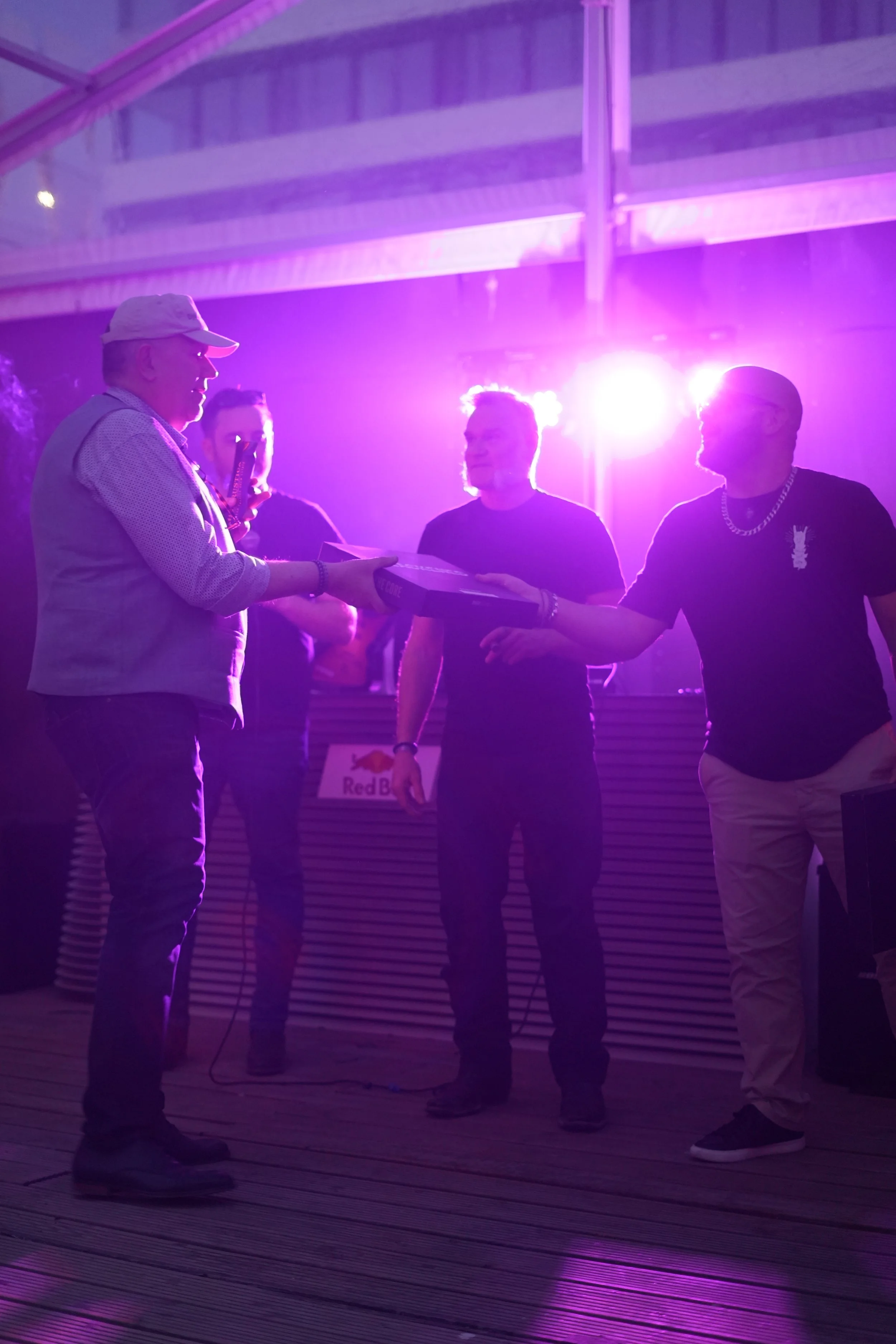 Four people standing in a dimly lit environment with purple lighting, with one person handing something to another.