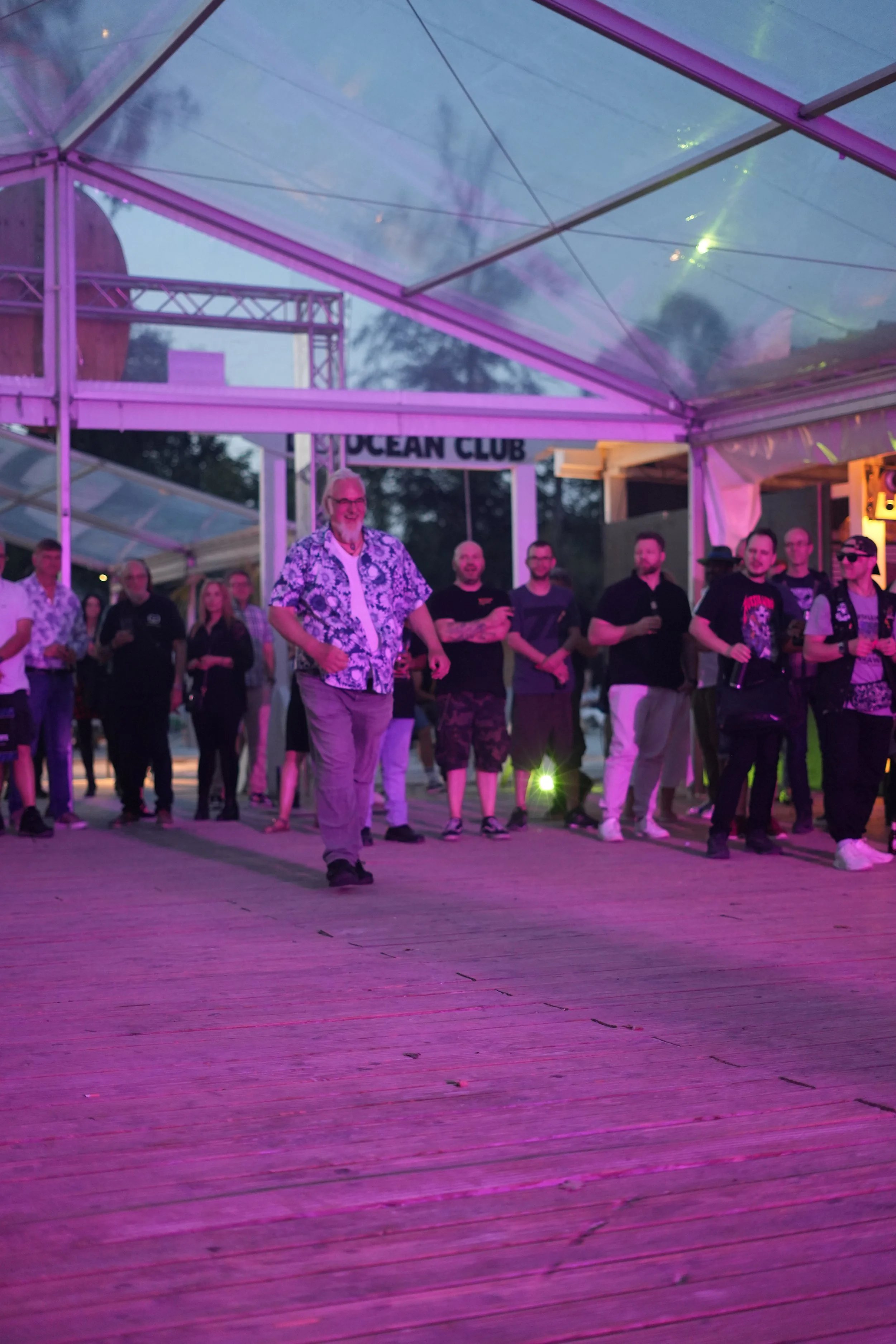 People dancing and socializing under a transparent tent with purple lighting at an outdoor event.