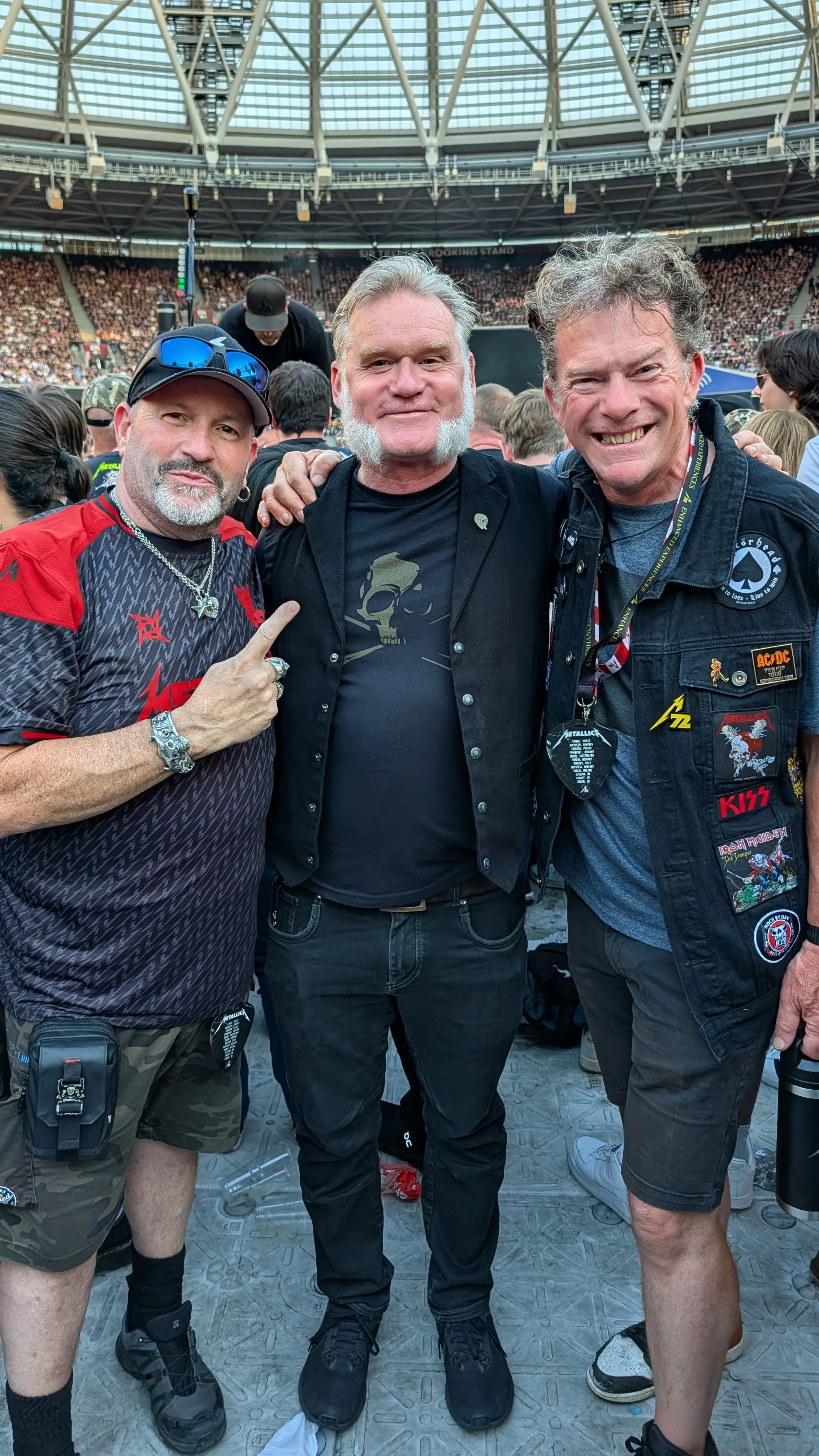 Three men standing together at a concert, with a stage and crowd in the background. The man on the left is pointing at the man in the middle. The man on the right is smiling widely. All are dressed casually with band patches and accessories.