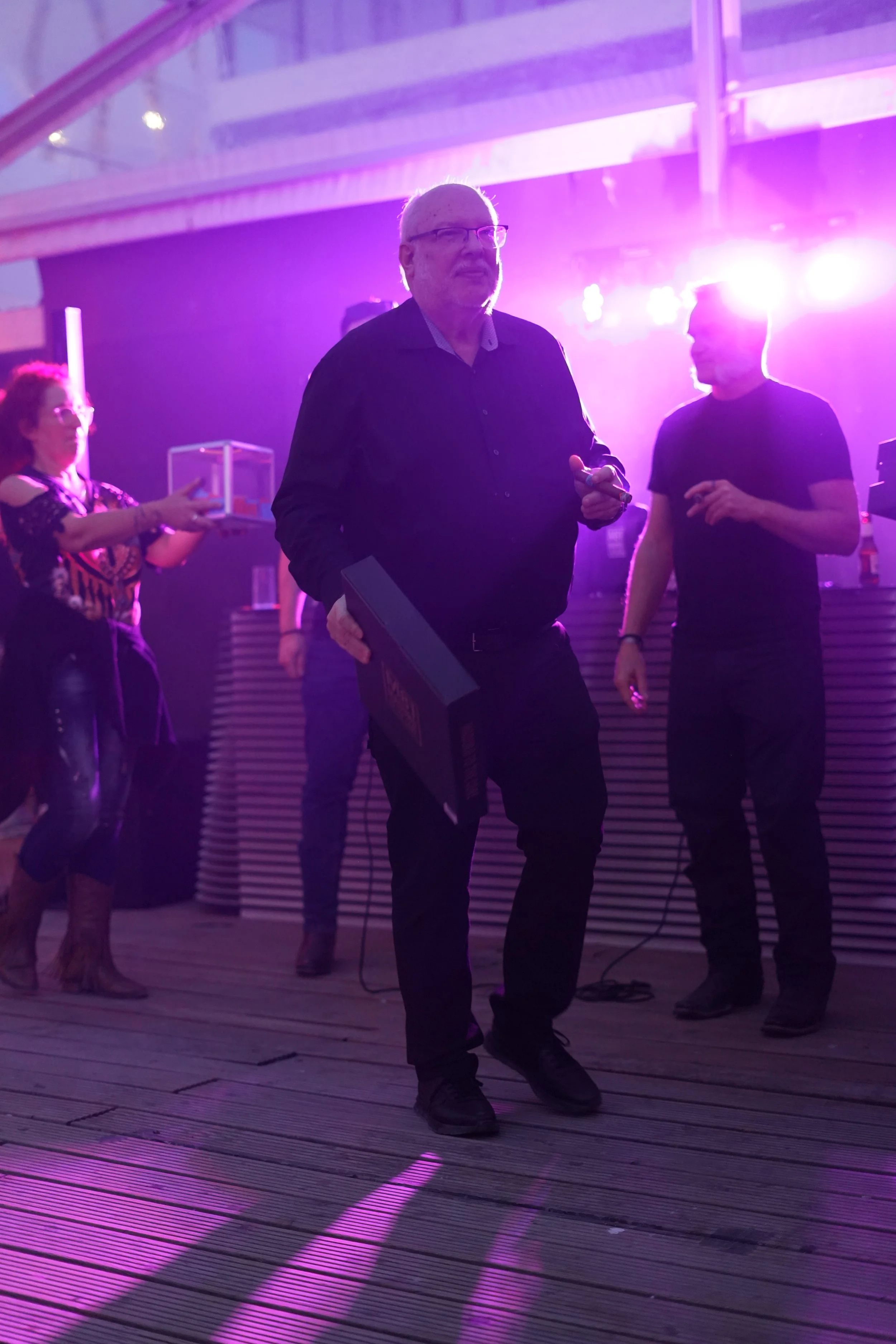 An older man with glasses and a beard, wearing a black shirt, is holding a book or DVD case and standing on a wooden floor illuminated by vibrant purple and pink stage lights, with other people in the background at an indoor event or party.