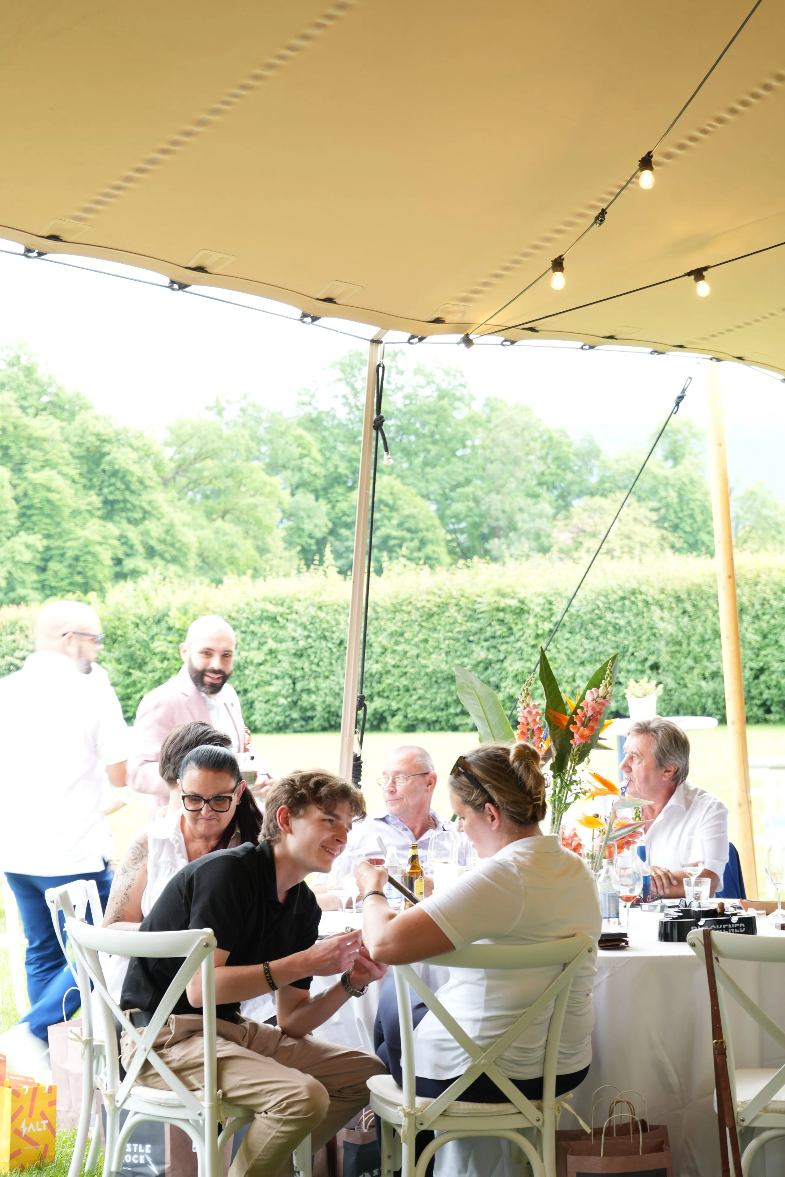 People gathered around a table at an outdoor event under a canopy with string lights, with trees and green landscape in the background.