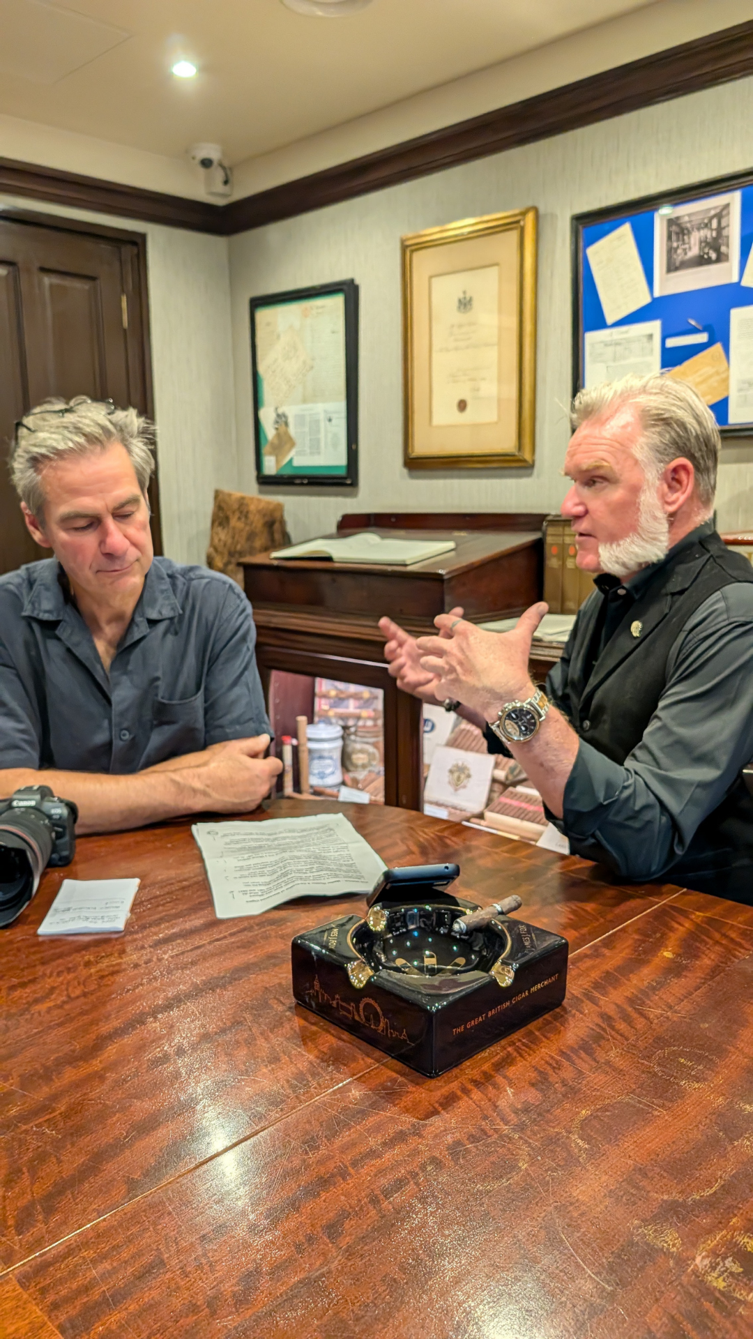 Two men having a conversation at a wooden table in an office or library setting. One man is speaking, and the other is listening with arms crossed. The table has a camera, papers, and a decorative cigar box.