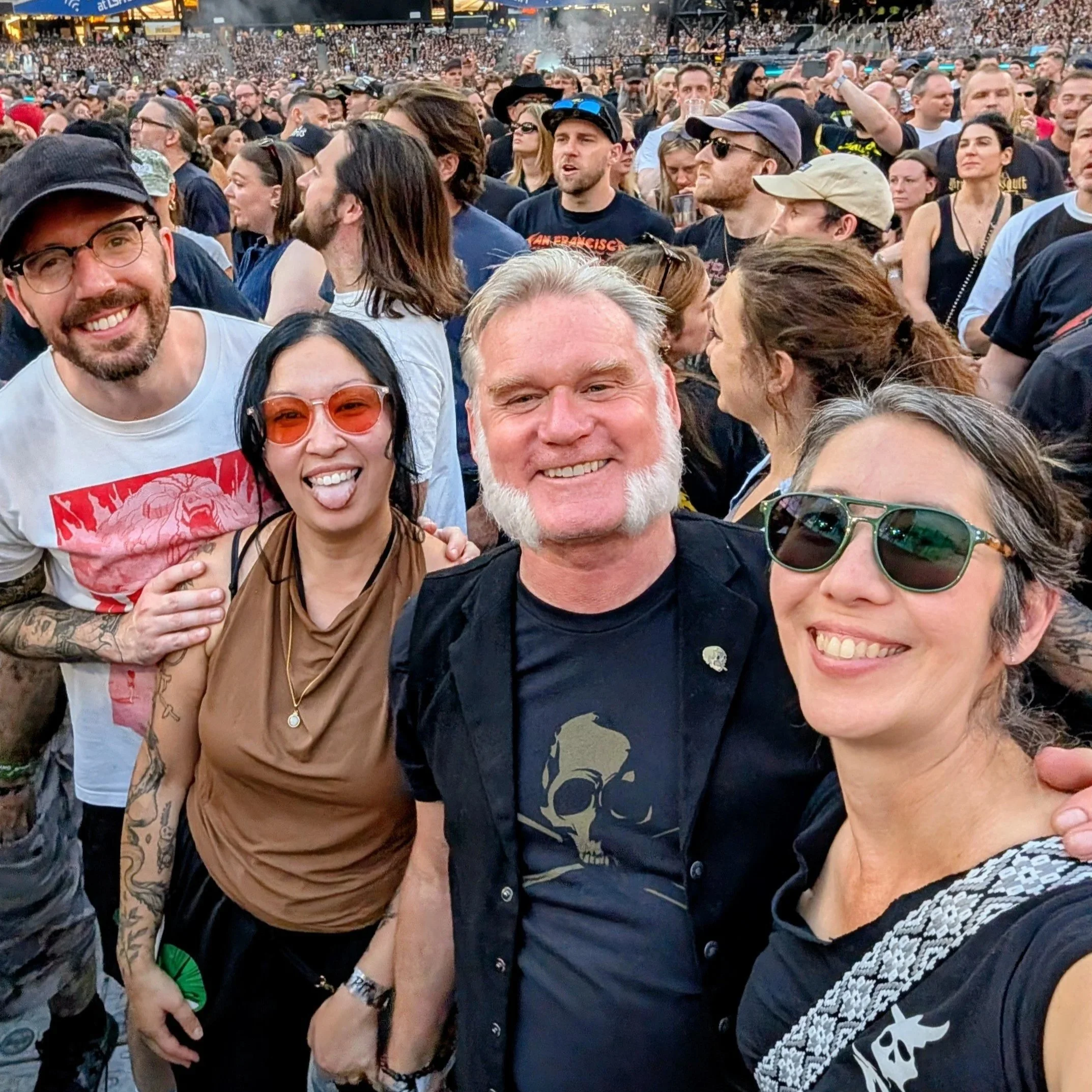 Crowd of people at a Metallica concert, with some smiling and posing for the photo, and others talking or enjoying the event.