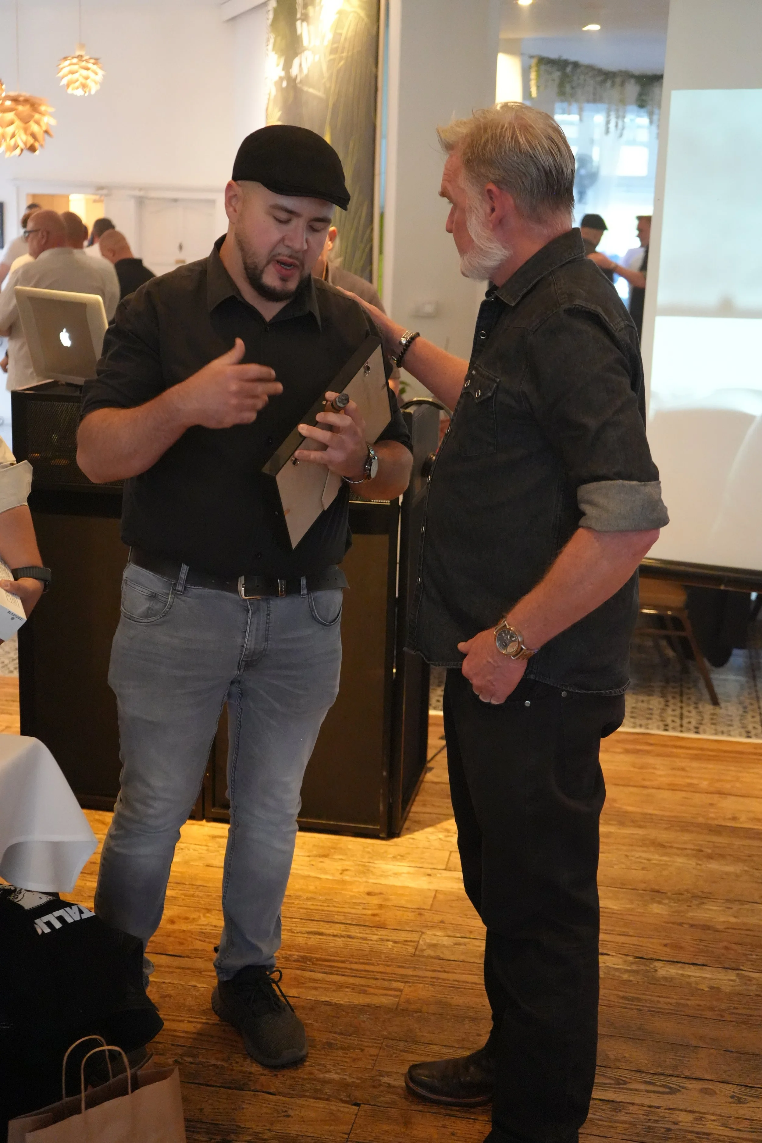 Two men are having a conversation in a social setting, with one appearing to show or explain something on a tablet to the other. They are standing on a wooden floor, with one man in a black shirt and gray jeans, and the other in a black denim shirt a