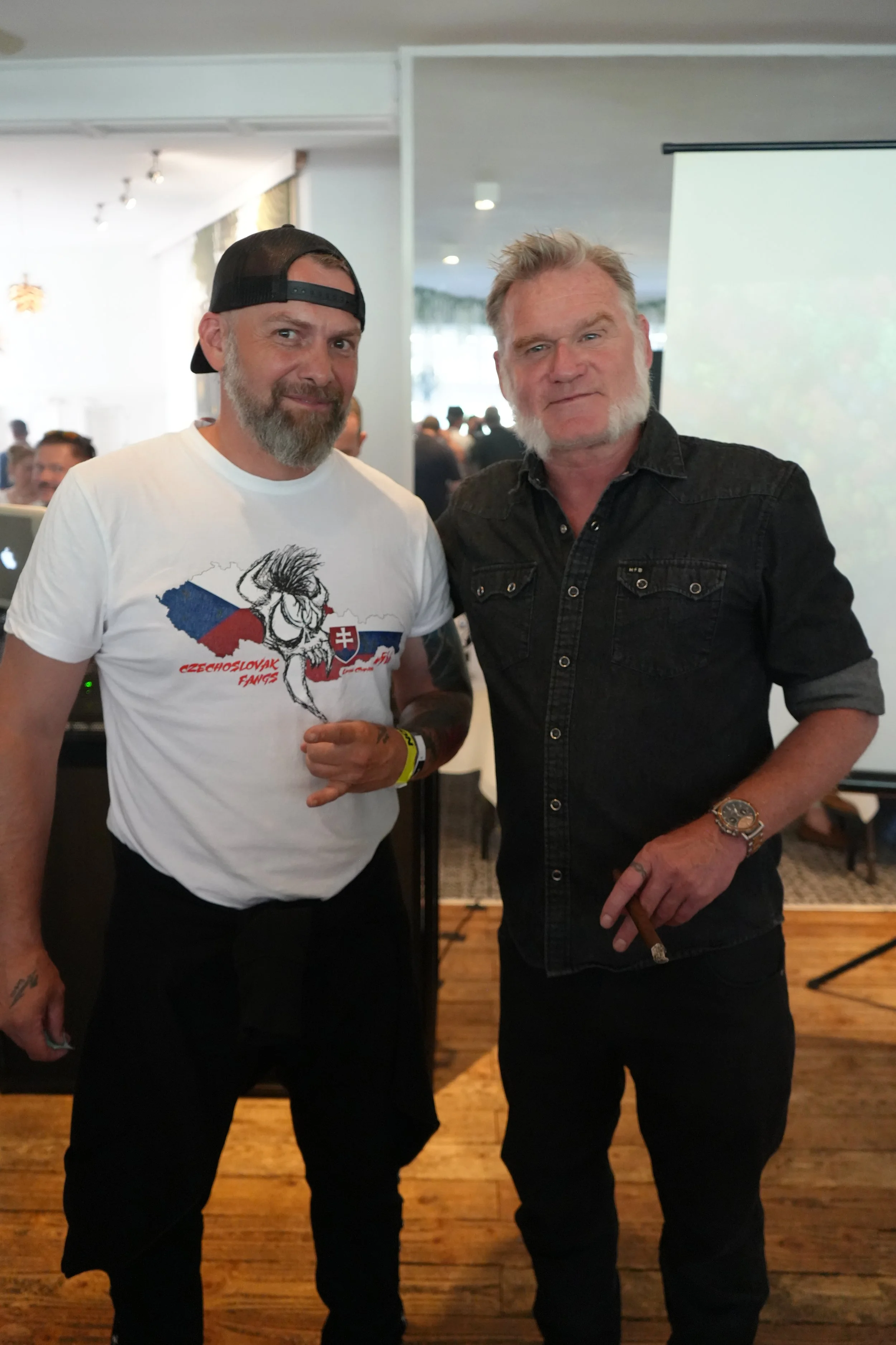 Two men standing indoors, one wearing a white t-shirt with a graphic of a creature and the words "Czechoslovak Fangs" and the other in a black button-up shirt, holding a cigar, both looking at the camera.