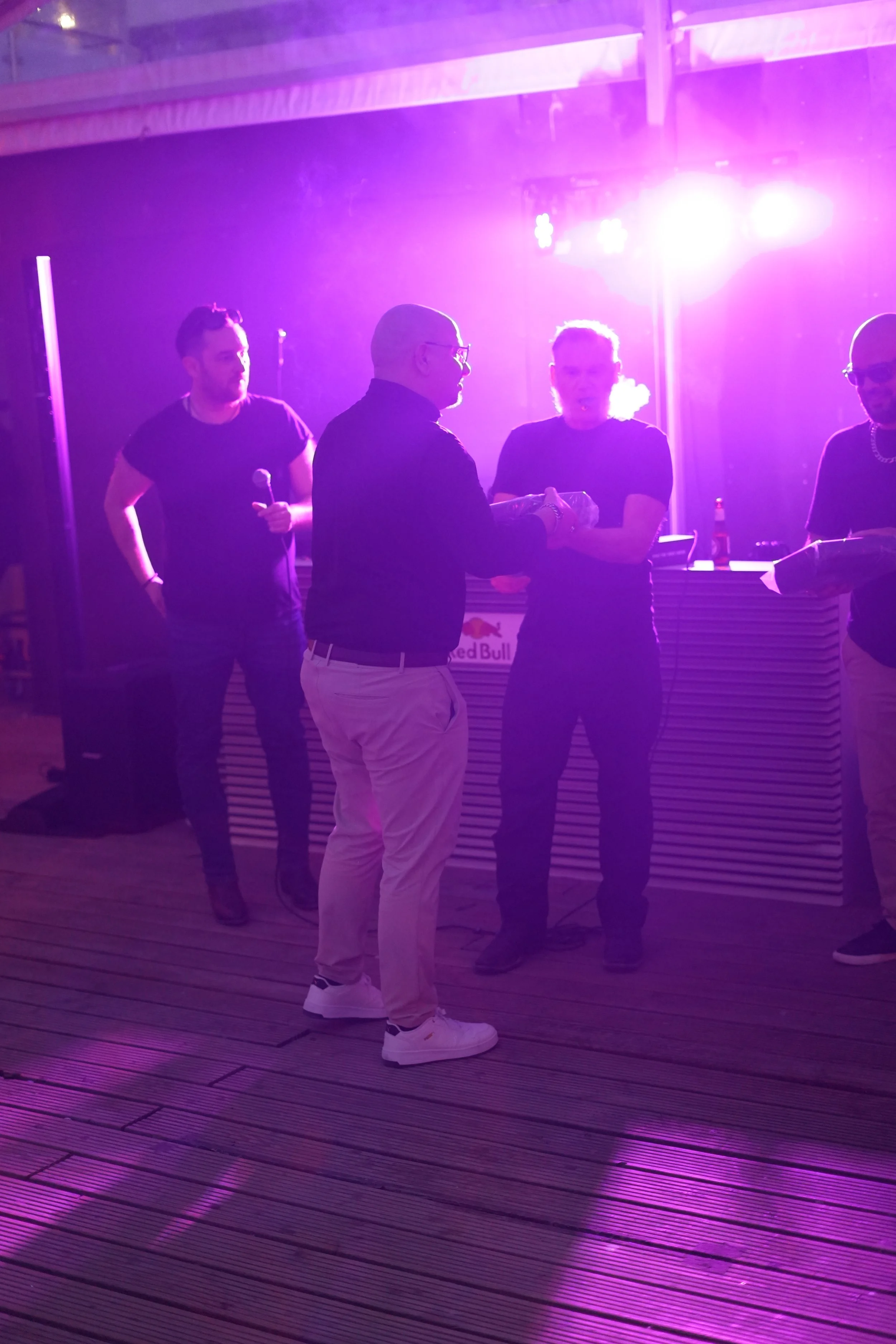 Four men are standing and conversing under purple and pink stage lighting, with a DJ booth; one man holds a microphone, the others hold drinks or papers.