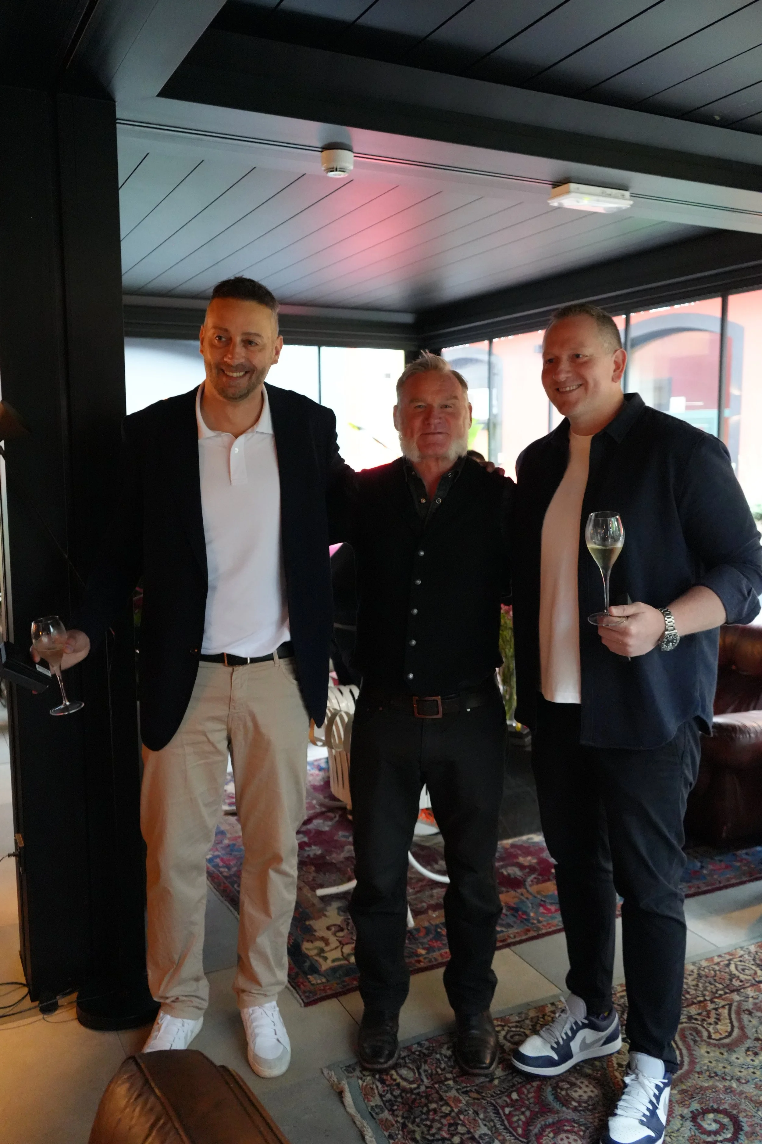 Three men standing together in a room, smiling for the camera. They are dressed in smart casual attire, holding glasses of champagne, with a patterned rug on the floor and large windows in the background.
