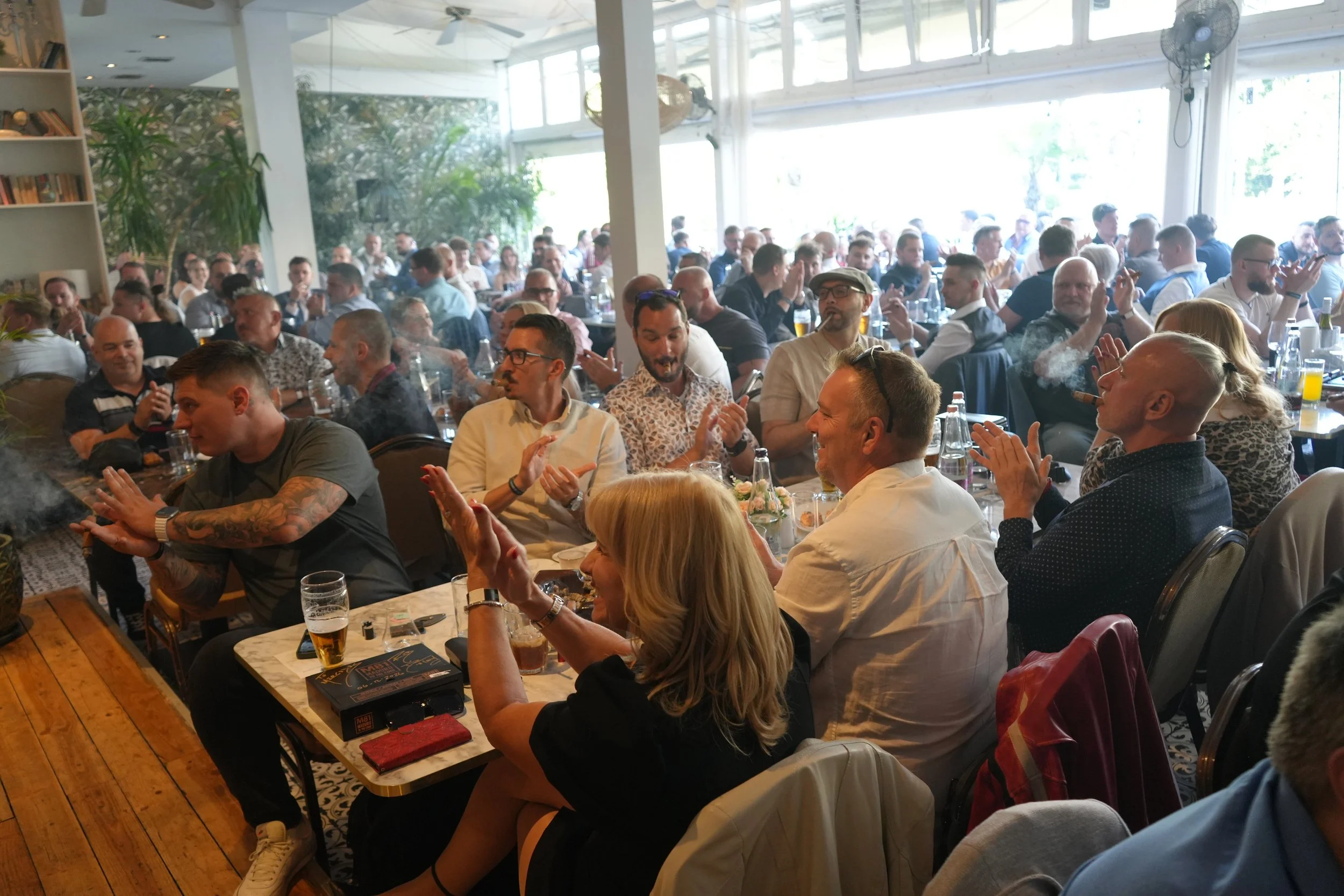 A large group of people gathered in a bright, indoor restaurant or event space, seated at tables and socializing.