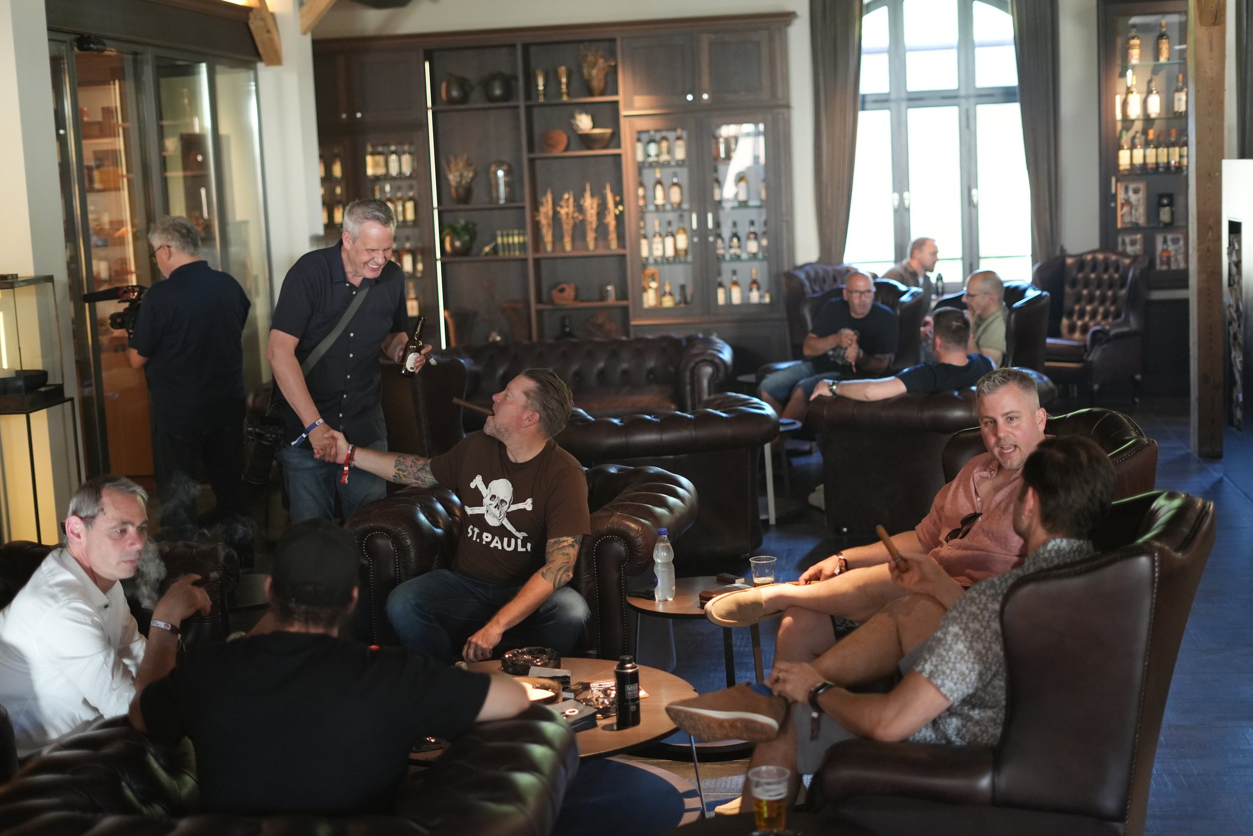 Group of men socializing and enjoying drinks in a cozy bar or lounge with large windows, wooden shelves, and leather furniture.