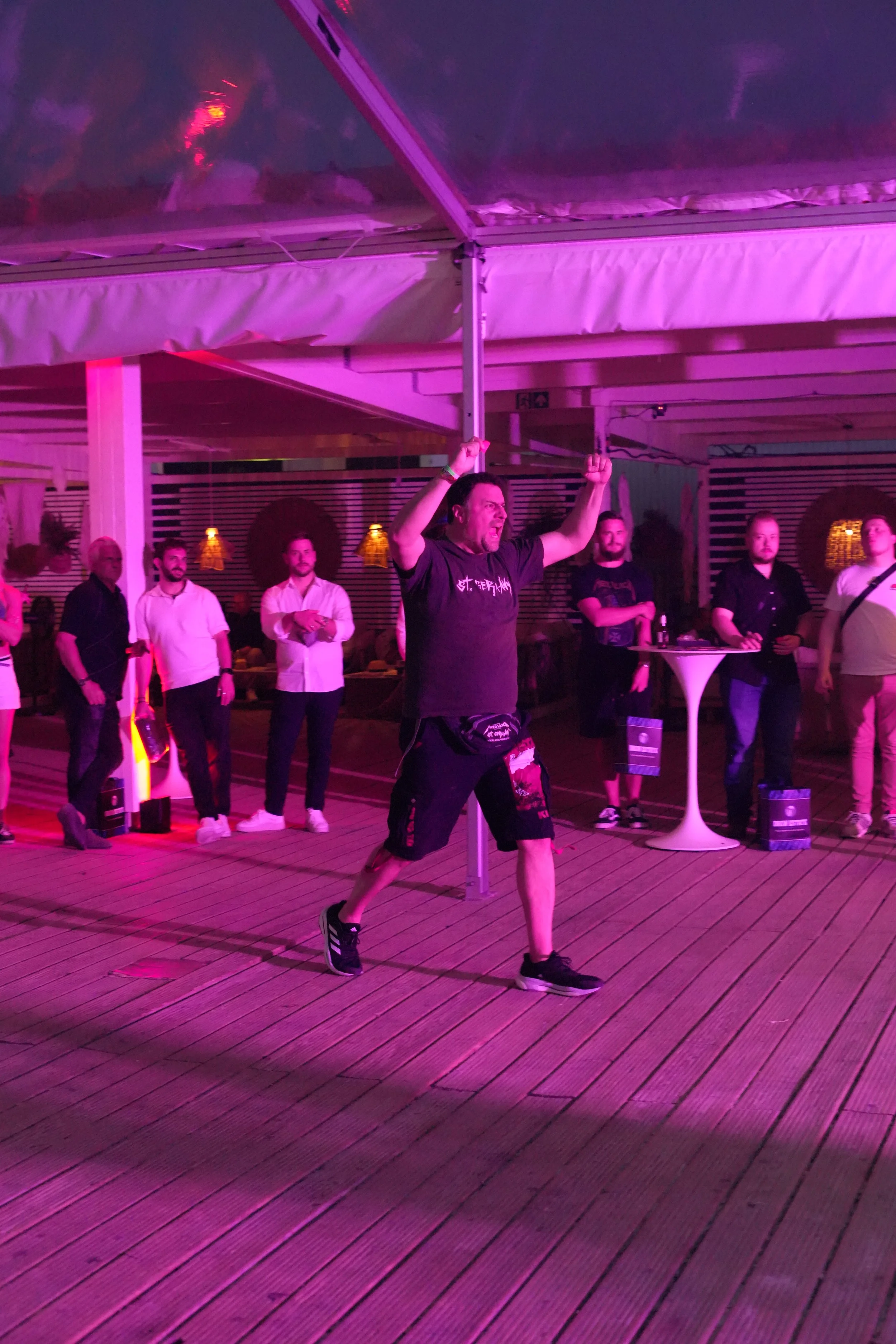 A man in a black t-shirt and shorts is dancing or performing with a pole in a dimly lit indoor setting with pink and purple lighting, and several people are standing around watching.