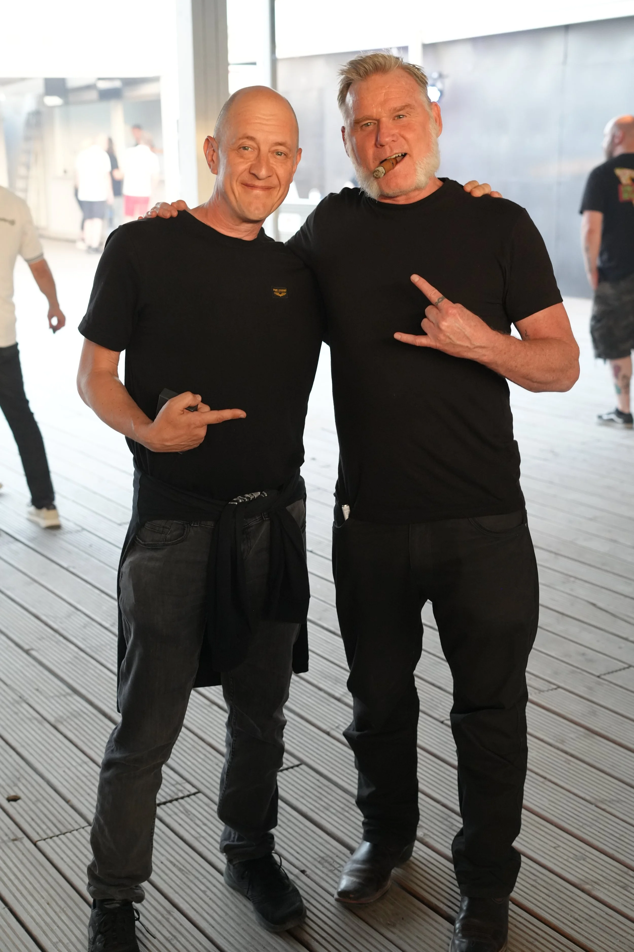 Two men standing together, smiling and posing, one of them has his arm around the other's shoulder, in an indoor setting with wooden flooring and blurred background of other people.