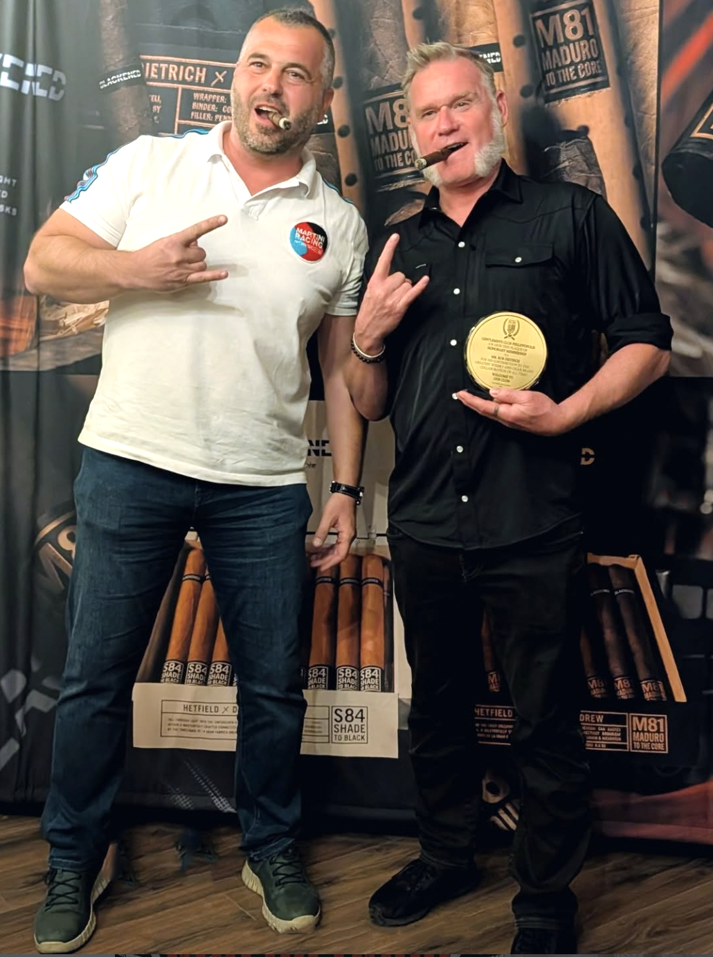 Two men standing together at an event, with cigars in their mouths. The man on the right is holding a gold-colored award plaque. Both men are making hand gestures, with the man on the left pointing at himself and the man on the right making a rock an