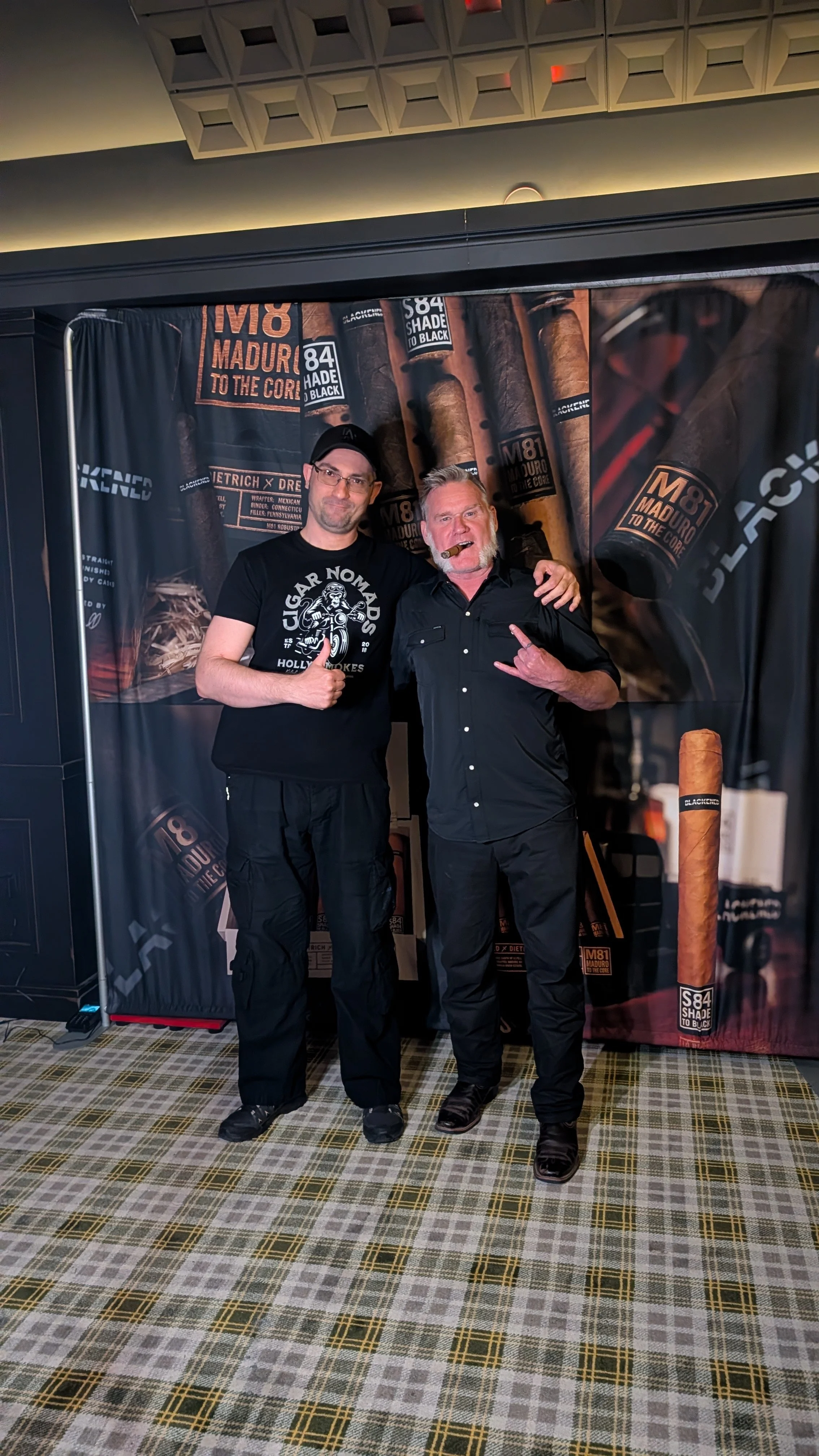 Two men standing together at a cigar event, with backdrops featuring cigar branding and images of cigars. One man is giving a thumbs-up, the other is making a 'rock on' hand gesture and smoking a cigar.