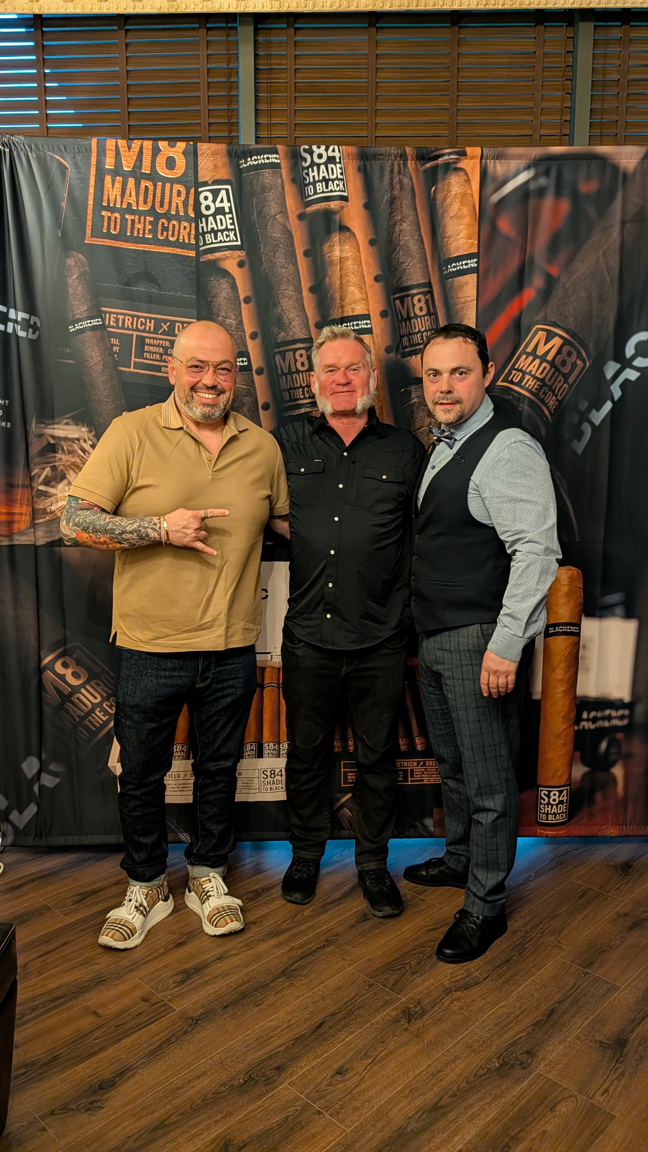 Three men standing in front of a backdrop with cigar images and text, smiling for the camera.
