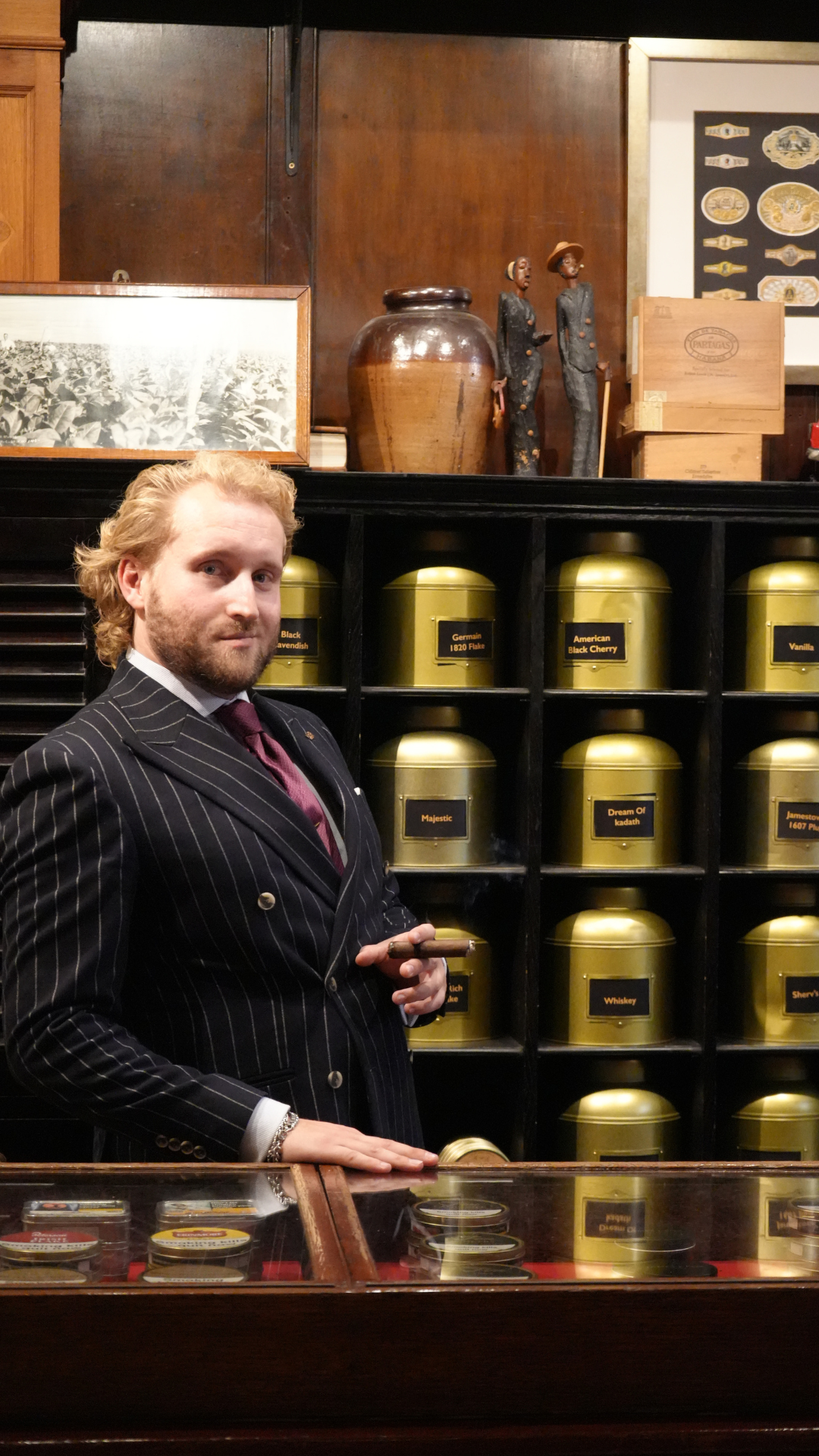 A man in a pinstripe suit holding a cigar in a vintage shop with gold tins of tea labeled with various flavors on the shelves behind him.