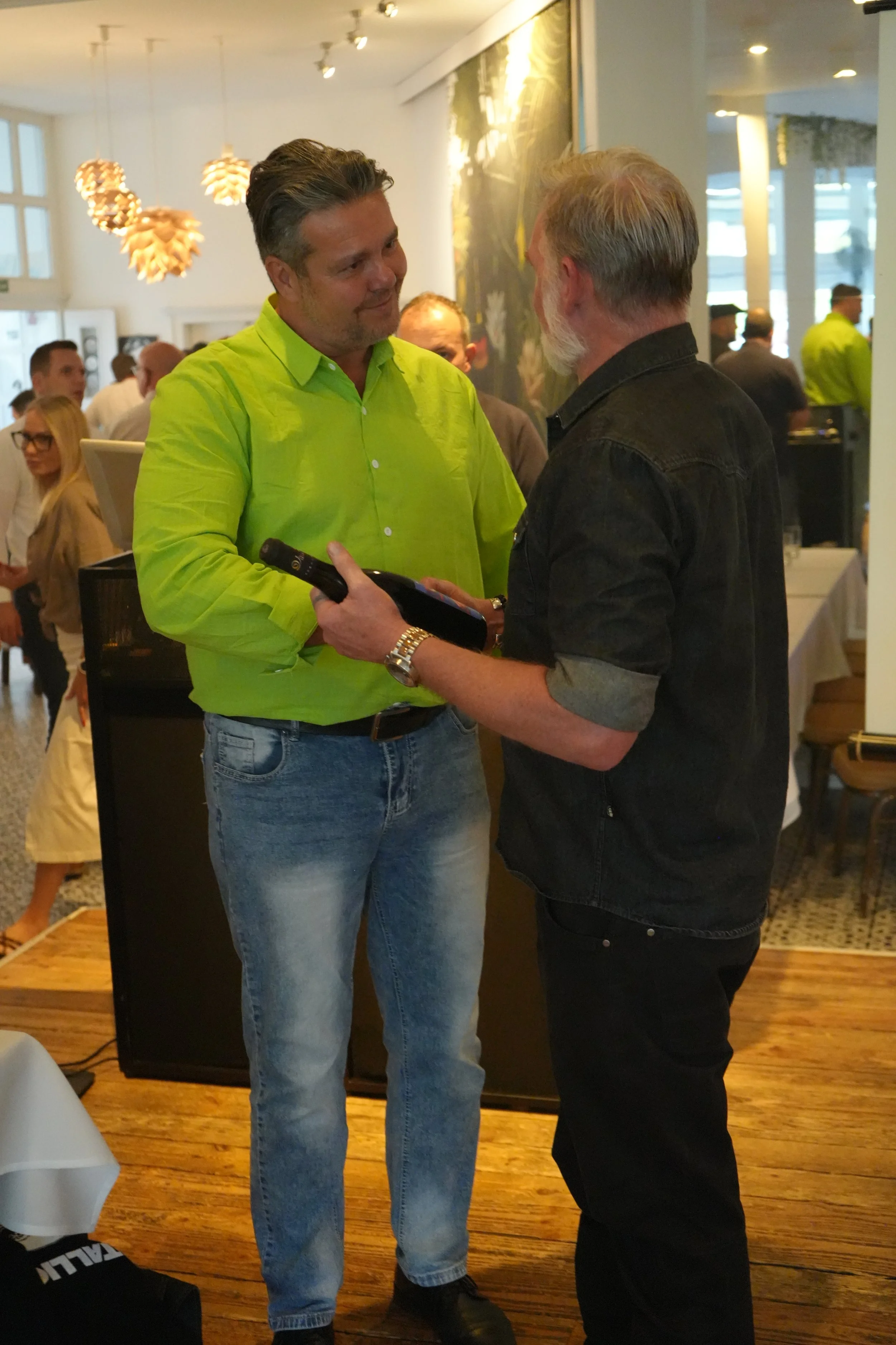 Two men are having a conversation at a social gathering or event inside a well-lit room. The man on the left is wearing a bright green shirt and jeans, holding a bottle, while the man on the right is dressed in a black shirt and dark pants. There are