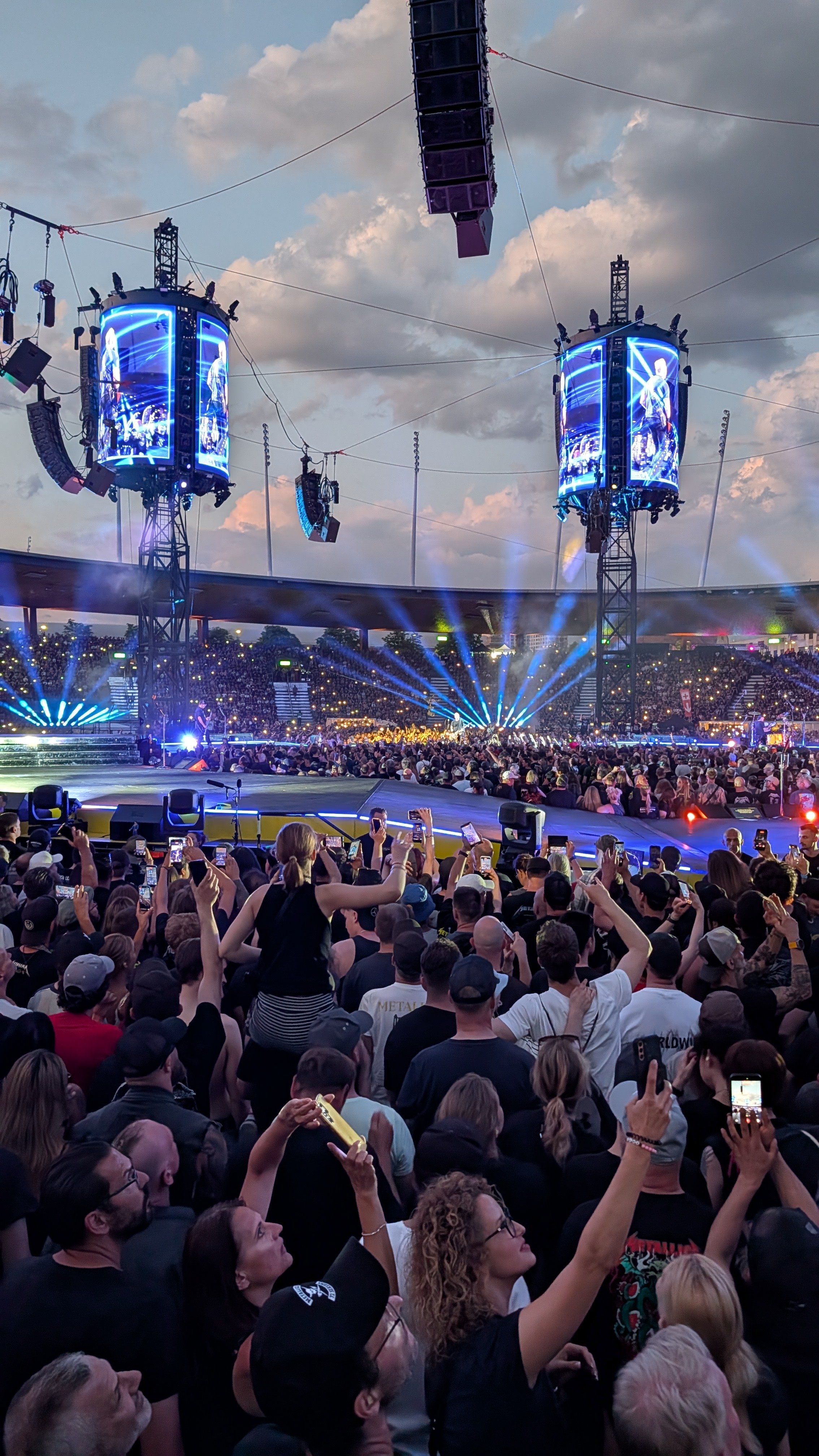 A crowded outdoor Metallica concert with people taking photos and videos of the stage, which has large screens and bright blue lights, under a cloudy sky.