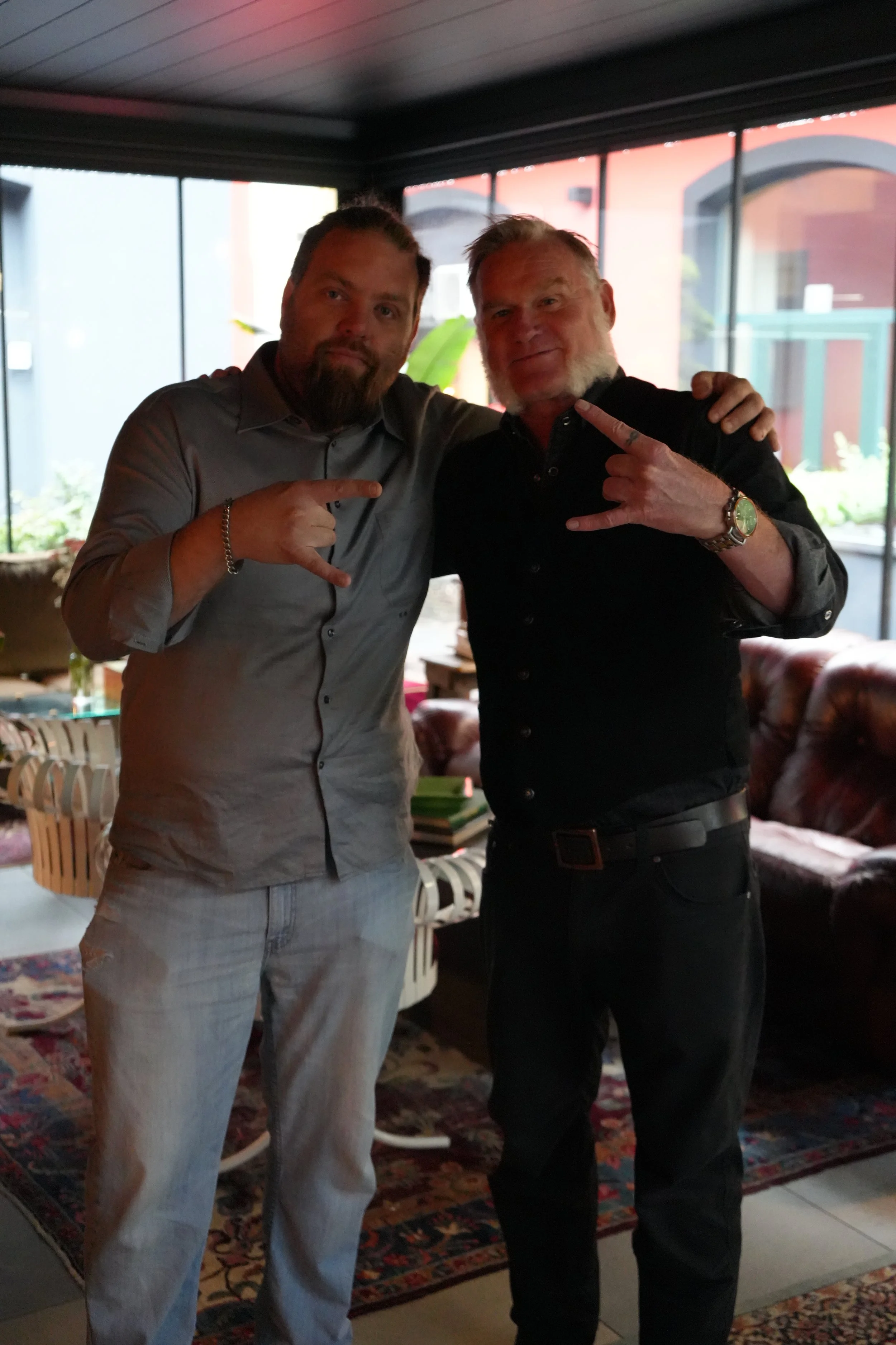 Two men standing indoors, pointing at each other, in casual attire. One is wearing a gray shirt and gray pants, and the other is wearing a black shirt and black pants. They are in a room with large windows, a colorful rug, and furniture including cha
