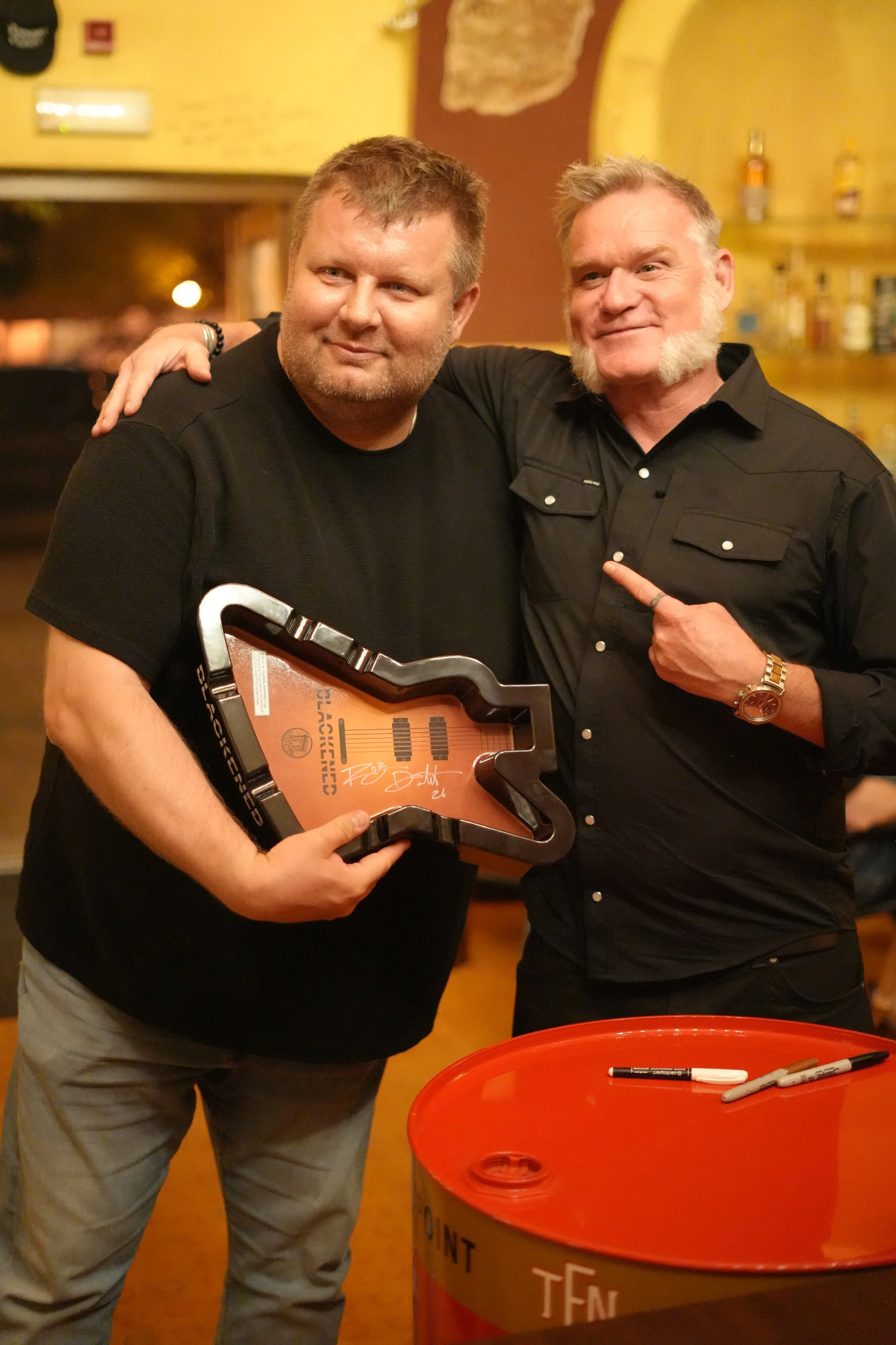 Two men smiling and posing for a photo in a bar or restaurant, one holding a guitar-shaped plaque. There are markers on a red barrel in front of them.